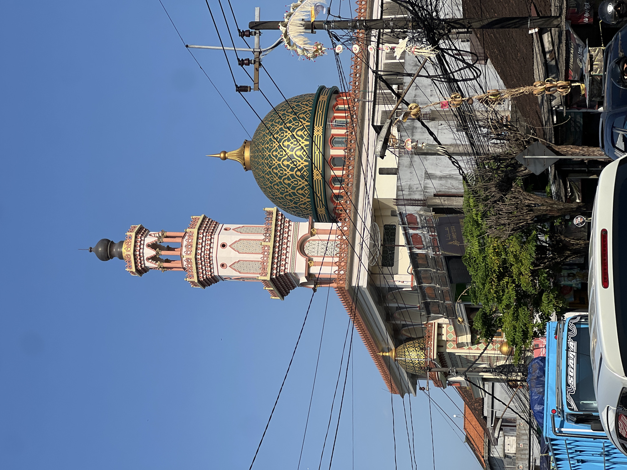 This image depicts a mosque in Kediri, Indonesia, under a clear blue sky. The mosque features a tall, multi-tiered minaret with pink and white accents, topped by a dark dome. A large, green dome with intricate gold patterns adorns the main structure, supported by red and white architectural elements. Several smaller golden domes are also visible. The mosque is partially obscured by a dense network of electrical wires and a utility pole with hanging decorations. In the foreground, a blue truck and the roof of a white car are visible, suggesting a street-level perspective in an urban environment. Adjacent buildings with tiled roofs and some trees add to the urban landscape. The overall scene captures a blend of religious architecture and the everyday infrastructure of a city.