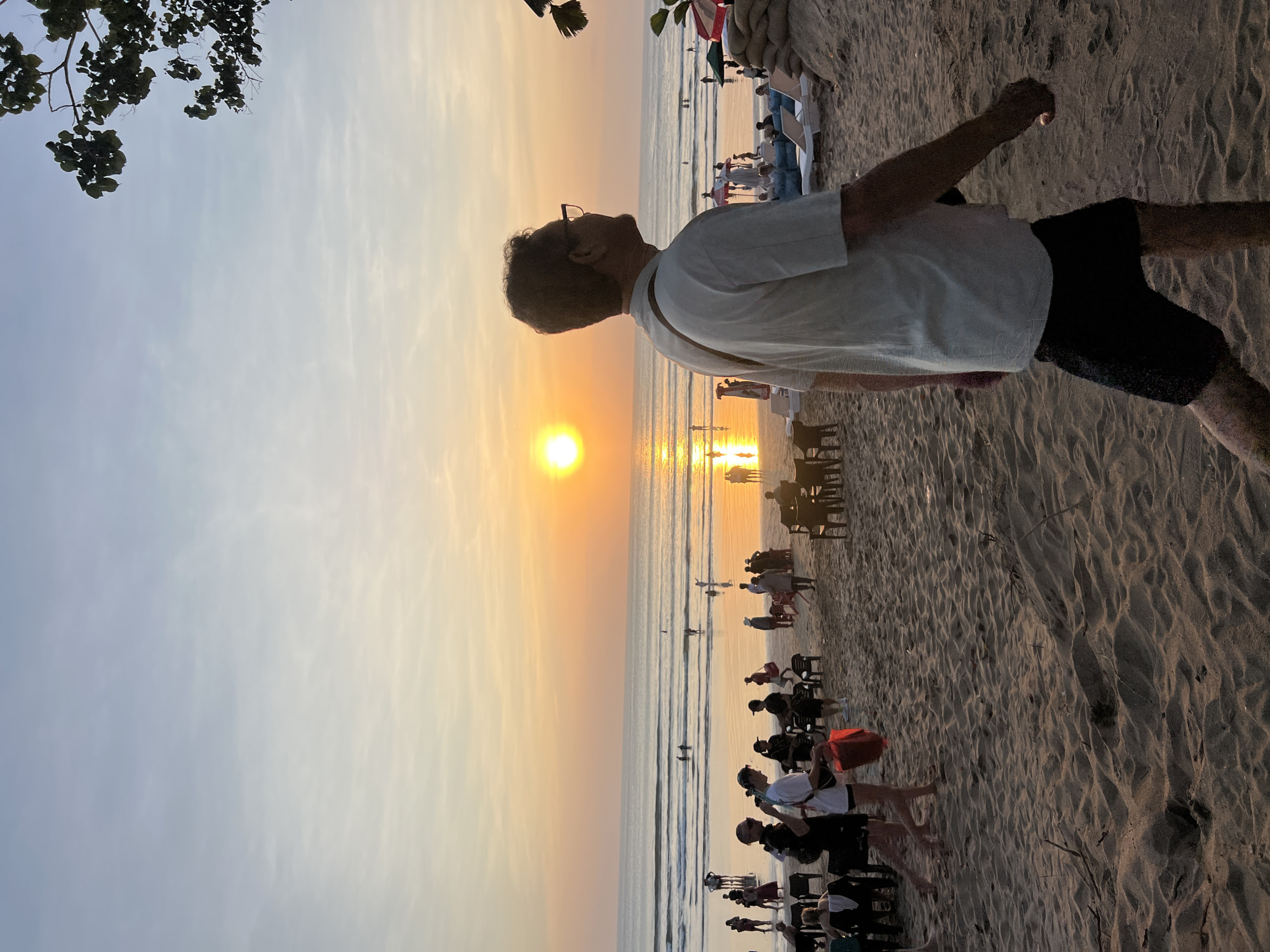 A medium shot captures a man in a white t-shirt and black shorts walking on a sandy beach in Kuta, Indonesia, during sunset. The sun, a bright orb, is low on the horizon, casting a warm golden glow across the sky and the ocean. Gentle waves roll onto the shore, where several people are visible, some wading in the water and others relaxing on lounge chairs. A few umbrellas and beach huts dot the scene, suggesting a tourist area. The sand is textured with ripples, indicating the movement of the tide. In the foreground, the man's profile is partially obscured as he looks out towards the sea. The overall atmosphere is serene and tranquil, characteristic of a beach at dusk.