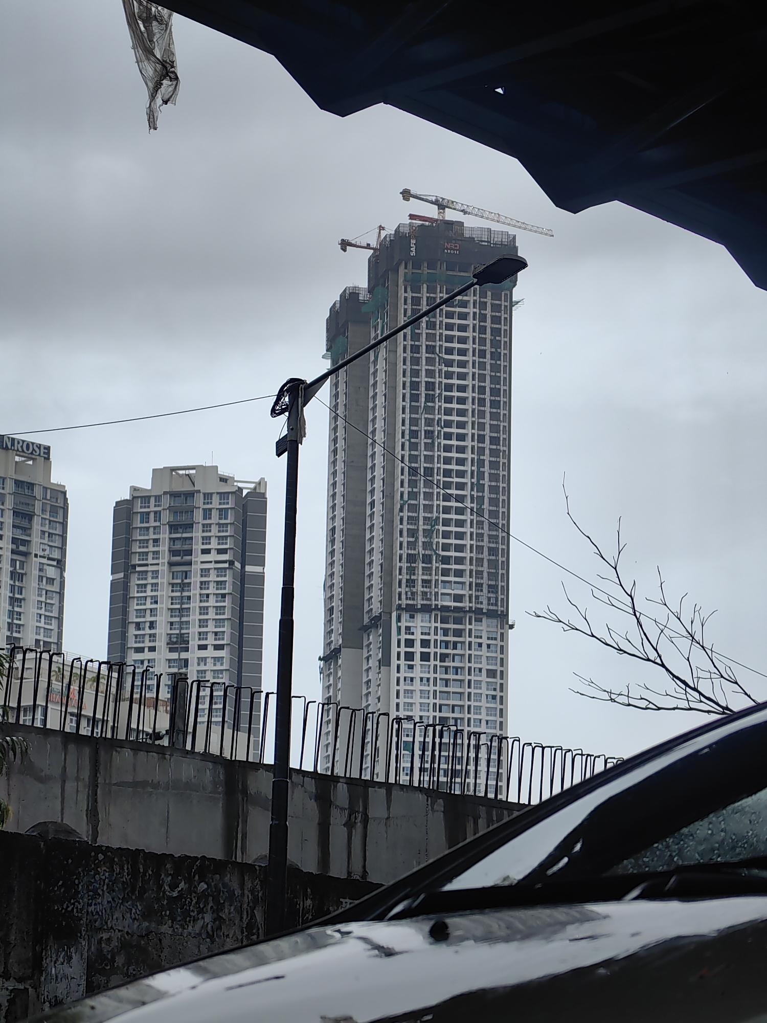 The image depicts a cloudy sky over a construction site in Mumbai, India. In the foreground, a black car is partially visible, with its windshield reflecting the overcast sky. Behind the car, a concrete wall with rebar sticking out of it lines the perimeter of the construction site. Several tall buildings are under construction, with cranes visible at the top of one of the tallest structures. One building in the middle ground is partially complete, showing rows of windows and balconies. Another building to the left, labeled "N. ROSE," appears to be near completion or already occupied. A street lamp stands in front of the buildings, with wires running across the frame. A piece of torn grey plastic hangs from an overhead structure in the upper left corner of the image. The overall scene suggests an ongoing urban development project in a dense city environment.