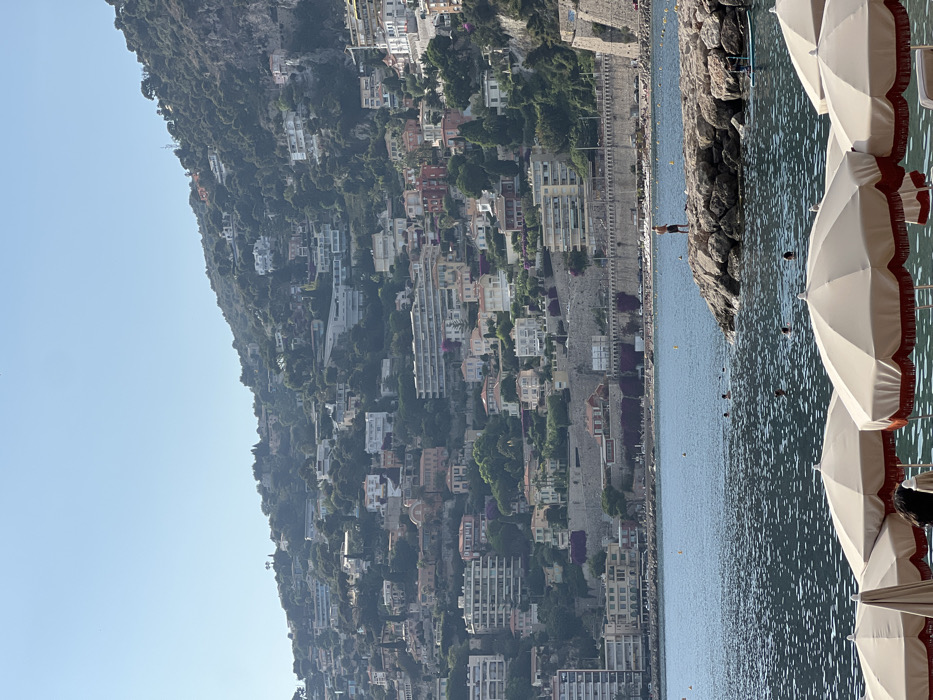 A hillside town descends toward the sea, densely packed with buildings among lush greenery. Below, a narrow road traces the coastline. The water reflects the sky's blue hue, dotted with buoys. A small rocky outcrop hosts a solitary figure. In the foreground, beach umbrellas stand in a row, their edges trimmed with a reddish-brown fringe. The umbrellas partially obscure the view.