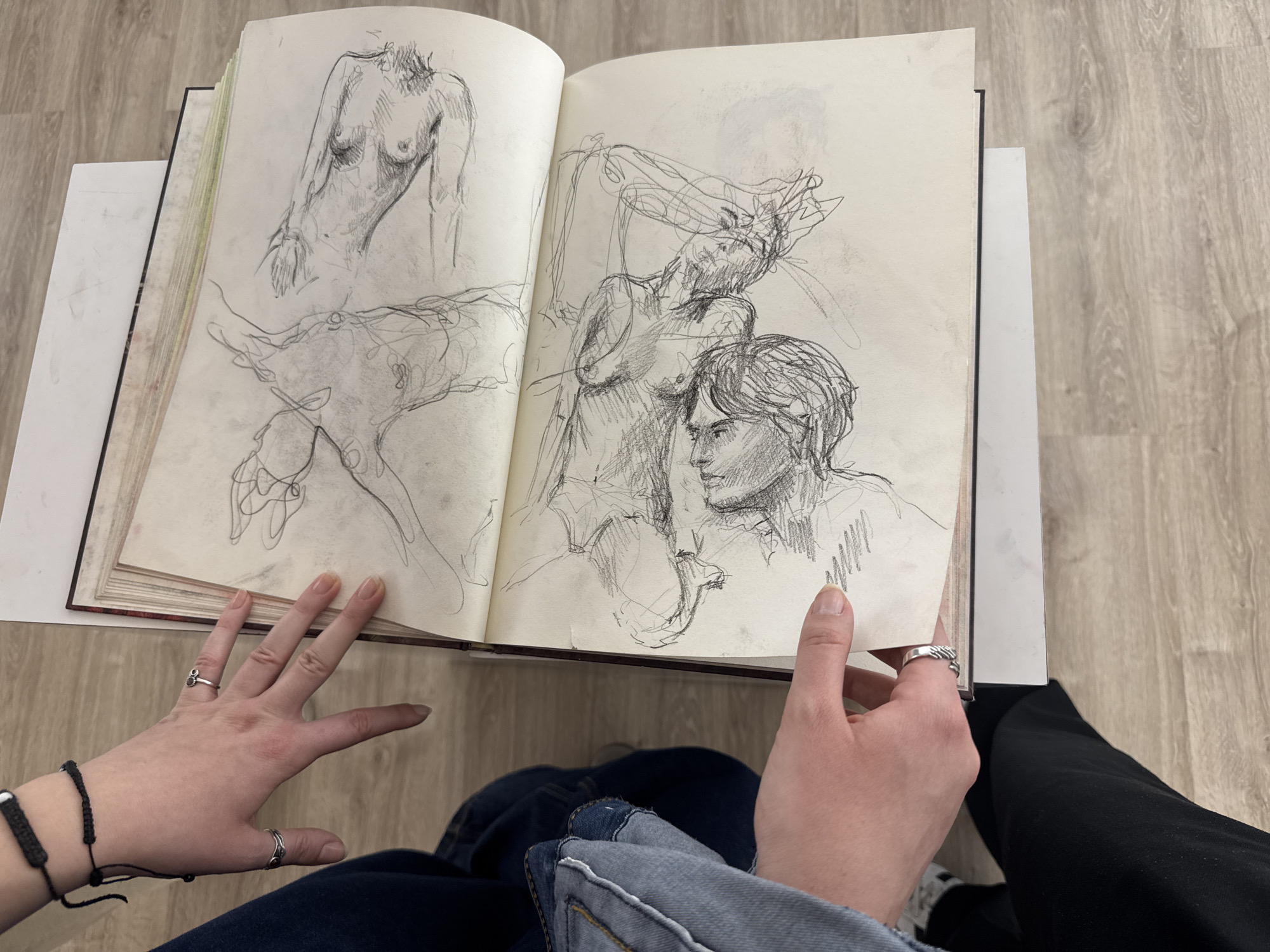 A person sits with an open sketchbook in their lap, displaying a two-page spread of figure drawings in pencil. The person is wearing dark jeans on their left leg and black pants on the right. A denim jacket is also visible. The left hand has a black bracelet and silver rings. The right hand also has a silver ring. The sketchbook rests on a small white table. The background shows a light-colored wood-patterned floor. There is no specific evidence to determine the location is in Yerevan, Armenia.