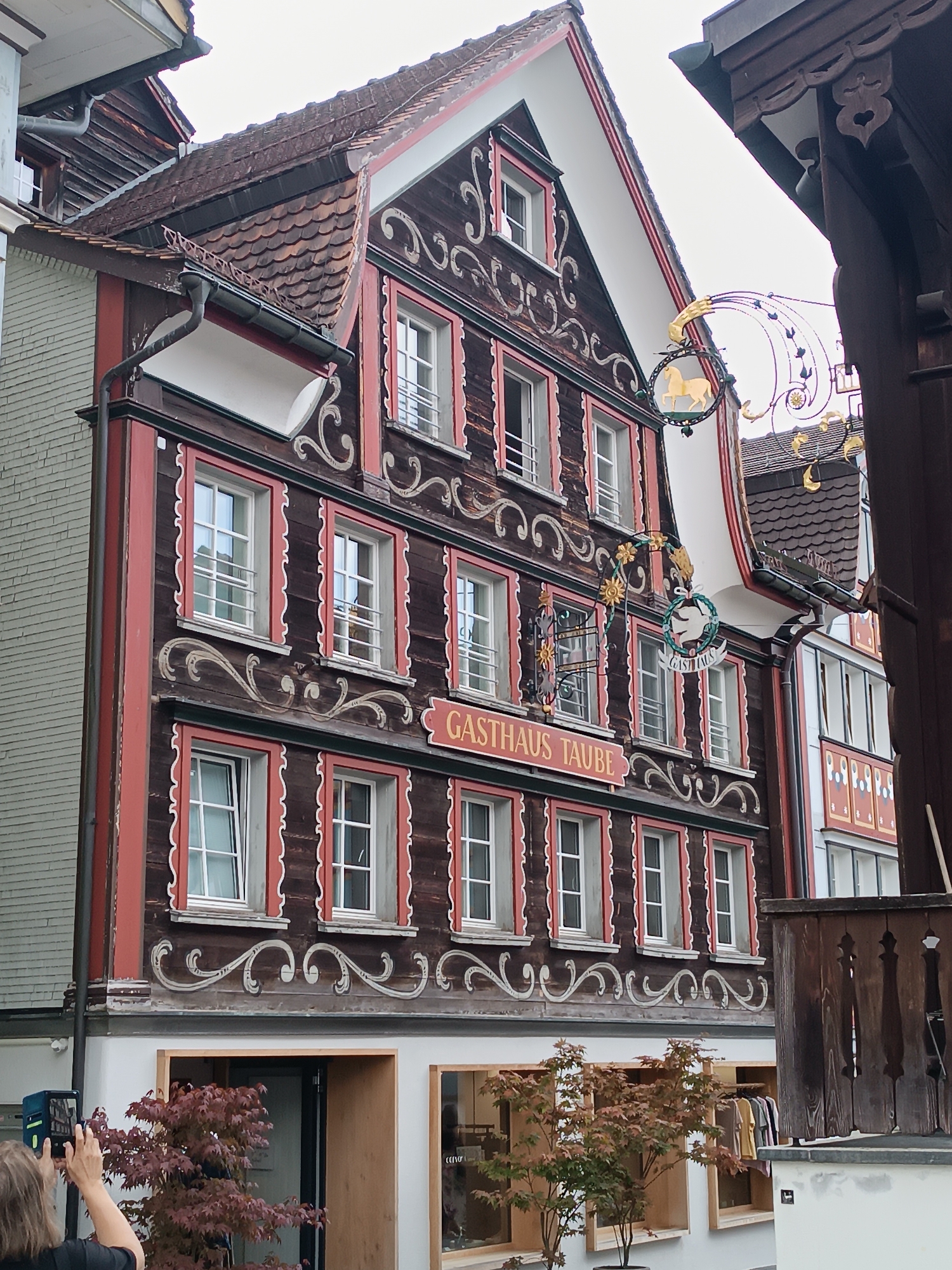 The image captures a facade of a building in Appenzell, Switzerland, likely a historic establishment with a traditional wooden exterior adorned with decorative carvings and red window frames. A prominent sign, "GASTHAUS TAUBE," is visible on the building's facade, indicating it is an inn or restaurant. Hanging signs with metallic decorations, including what appears to be a golden horse and a circular sign with "GASTHAUS" and a dove emblem, are suspended in front of the building. The building features multiple windows with metal grates, and the roof is tiled.
In the foreground, a person is partially visible on the left, holding a smartphone to take a photo of the building. A small tree with reddish-brown leaves stands in front of the ground floor entrance. The environment suggests an urban setting with other buildings visible, including one with a light-colored, horizontally ribbed facade to the left and another with colorful decorative panels to the right. The sky is overcast. The overall impression is one of traditional Swiss architecture in a town or village setting.