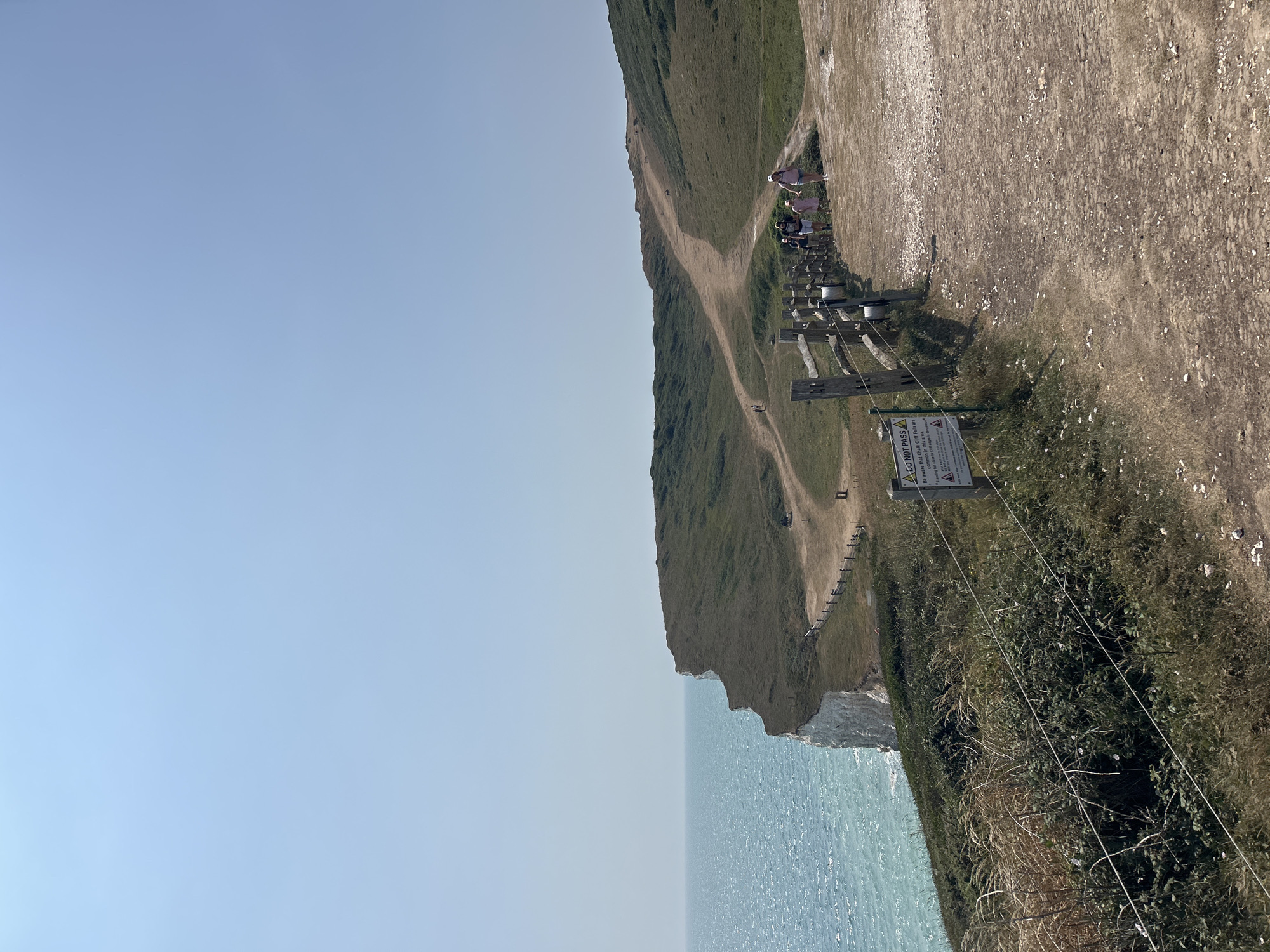 The image shows a section of the Seven Sisters cliffs near Seaford, United Kingdom. A dirt path winds along the cliff edge, with a wooden fence bordering it. Several people are walking on the path. The sea is to the left of the frame, and rolling green hills stretch into the distance. A "DO NOT PASS" sign is visible on a post near the fence, warning of the dangers of chalk cliff falls and unstable edges. The sky is clear and blue.