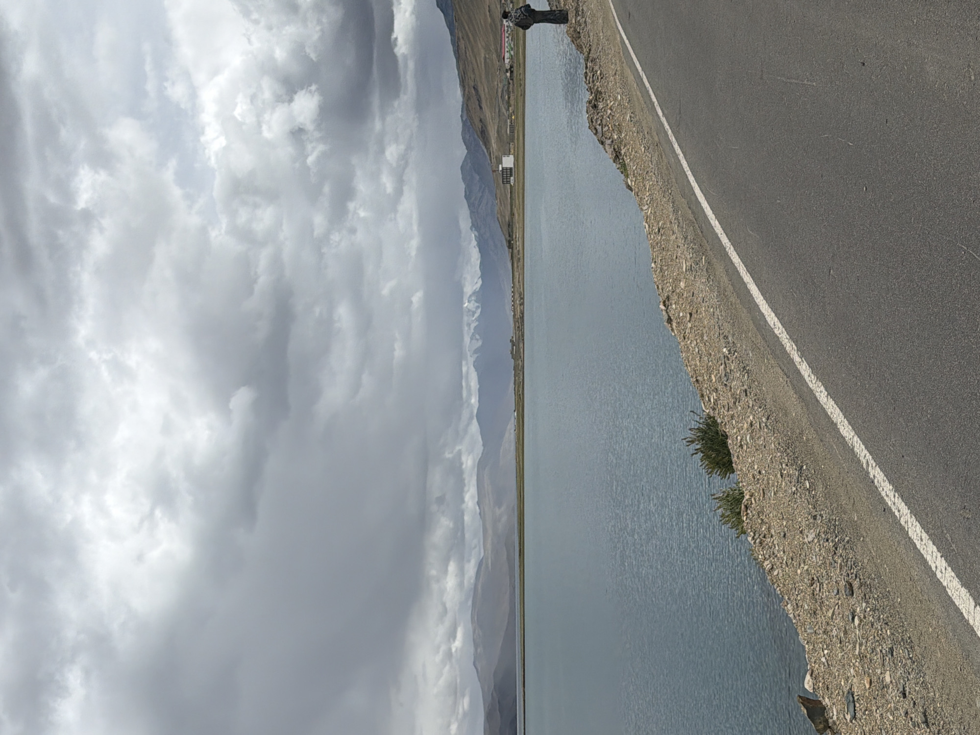 A lone individual stands on the side of a paved road, looking out over a large, calm body of water. The road curves to the right, with a gravelly shoulder that slopes down to the water's edge. The water is a deep blue and stretches towards a distant shoreline with rolling, barren hills. In the far distance, snow-capped mountains are visible under a dramatic, cloudy sky. The person is wearing dark clothing and a patterned shawl or backpack. The scene appears to be located in the high-altitude desert region of Karzok, India, near the Tso Moriri lake. The overall atmosphere is one of solitude and the vastness of nature.