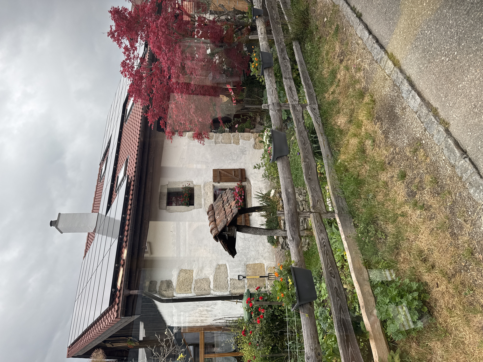 This is a frontal view of a stone and stucco house with a red tile roof, located in Petit-Val, Switzerland. The house has several solar panels installed on the roof, a prominent white chimney, and two windows with wooden shutters and flower boxes. In front of the house, a rustic wooden fence spans the width of the image, with several dark grey planters holding flowers attached to its top rail. Behind the fence, a vibrant red Japanese maple tree is visible, its leaves contrasting with the more muted tones of the house and vegetation. The surrounding environment includes a grassy yard, a paved pathway to the right of the fence, and a green garbage bin and a garden tool near the left side of the house. The sky is overcast.