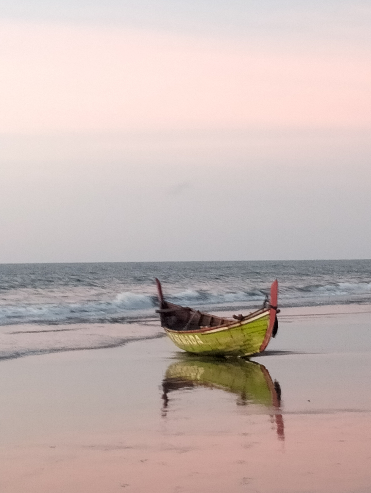 An eye-level, medium shot captures a solitary green and yellow fishing boat resting on the wet sand of a beach. The boat is oriented diagonally from the lower left to the upper right of the frame. Its hull is predominantly green with a yellow stripe running along the top edge. A red rudder or decorative piece juts upwards from the stern of the boat. The boat is positioned near the water's edge, with gentle waves lapping at the sand and creating a soft, white foam.
The sea stretches across the middle ground of the image, appearing calm and slightly choppy with visible waves. The horizon line is clearly defined, separating the sea from a vast, pale sky that transitions from a soft pinkish hue near the horizon to a muted gray at the top of the frame. The sky suggests either dawn or dusk.
The wet sand in the foreground acts as a mirror, reflecting the boat and the sky in a distorted, softened manner. The reflection is smooth, with hints of pink and yellow from the boat and sky. The overall lighting is soft and diffused, characteristic of twilight. The image appears to be captured in Uleekareung, Indonesia, a location known for its coastal scenery. There are no people visible in the scene.