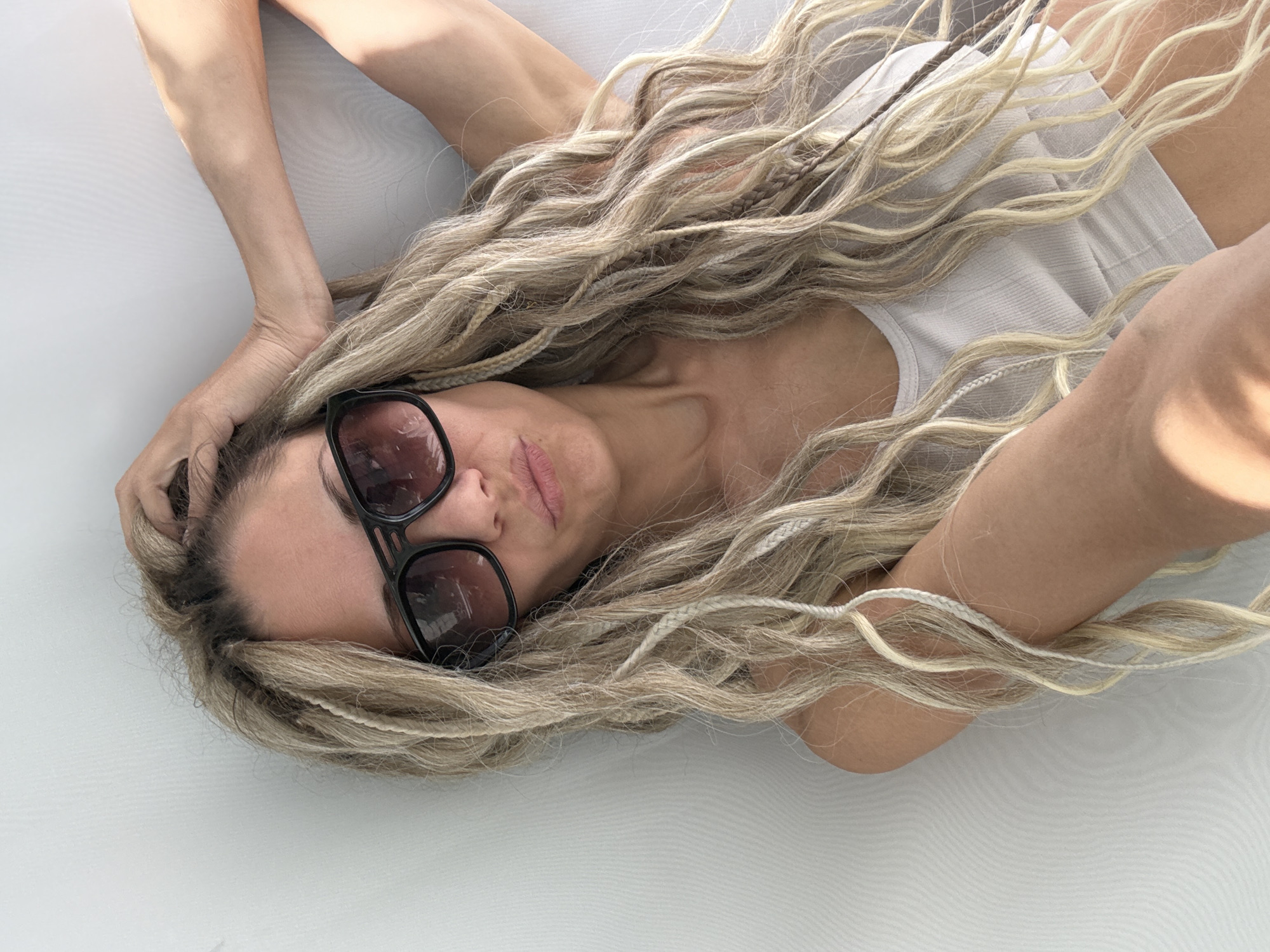 A person, likely female, is shown lying down from a high-angle perspective, with her body oriented horizontally across the frame. Her head is positioned towards the center-left, with her long, blonde hair fanning out around her. The hair features a mix of wavy strands and thin braids or dreadlocks. She wears dark-framed sunglasses that cover her eyes, and her lips are slightly parted, suggesting a relaxed or pensive expression.
The individual is wearing a light-colored, possibly white or off-white, ribbed tank top or camisole. Her arms are bent, with one hand resting near her head, partially obscured by her hair. Her skin appears tanned, and there are prominent shadows cast across her body and hair, indicating bright, direct lighting, likely from the sun. The background is a plain, light-colored surface, possibly a bed, floor, or outdoor lounge area. The overall scene conveys a relaxed, daytime atmosphere. There is no visible text in the image. The image is contextualized as being taken in Rusanovo, Russia.