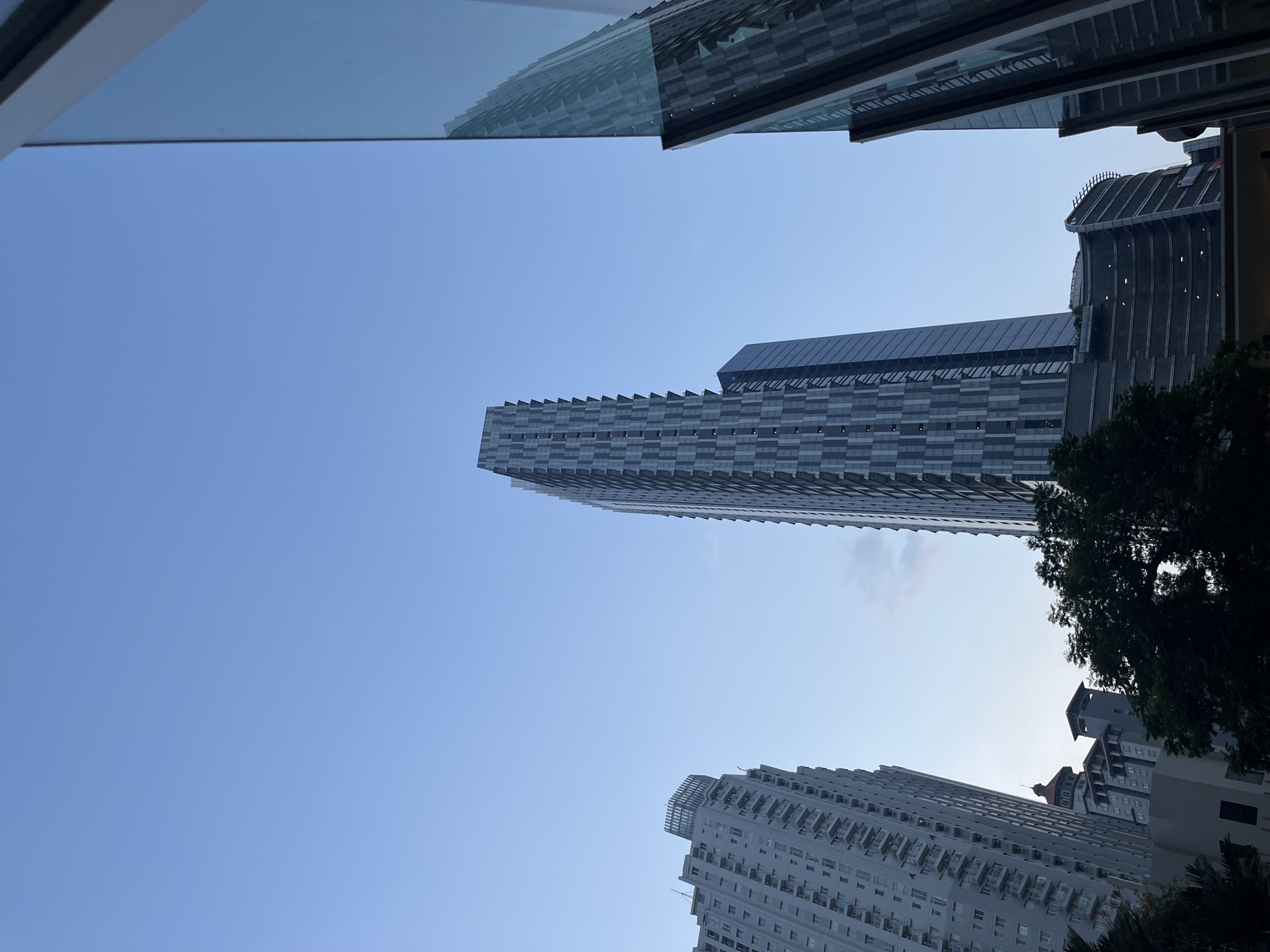 The image shows a low-angle view of several modern skyscrapers against a clear blue sky. In the foreground, on the right, a tall, sleek building with a glass facade reflects the sky and other buildings. To the left, two other skyscrapers are visible. One is a slender tower with a patterned facade, and another is a wider building with balconies. In the lower part of the image, some trees and the top of a lower building with distinct architectural features, possibly a dome and tiered roofs, are visible. The scene appears to be in an urban environment, likely a business or residential district in West Jakarta, Indonesia, given the architectural style and the clear, bright sky characteristic of the region. There are no people or animals visible in the frame.