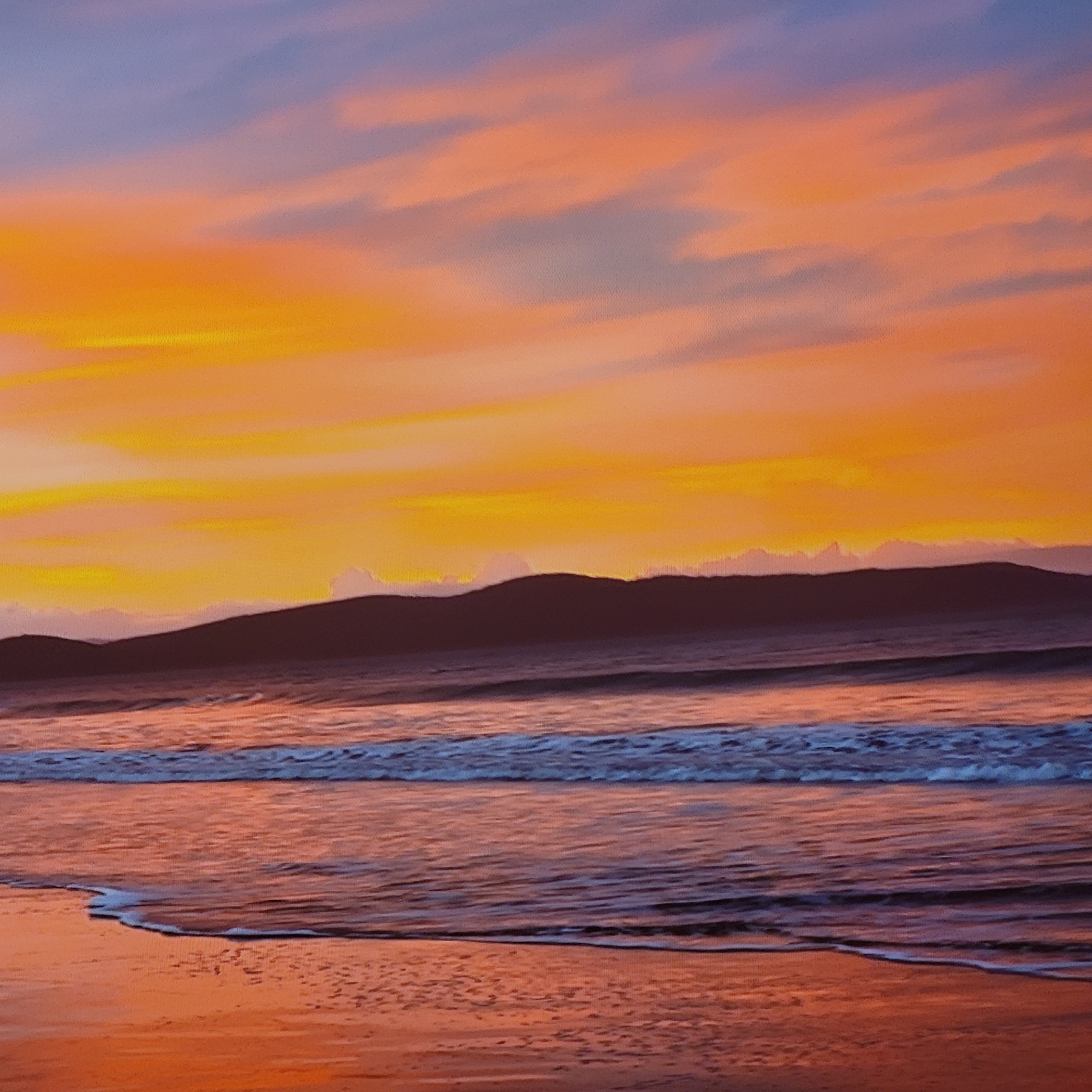 The image depicts a scenic sunset view of a beach, not located in Yerevan, Armenia, as the city is landlocked. The sky is a blend of orange, yellow, and hints of purple, with soft clouds. A dark silhouette of mountains or hills is visible in the middle ground. The ocean has gentle waves with a white foamy crest. The beach itself is a reddish-orange hue, reflecting the colors of the sunset. There are no people visible.