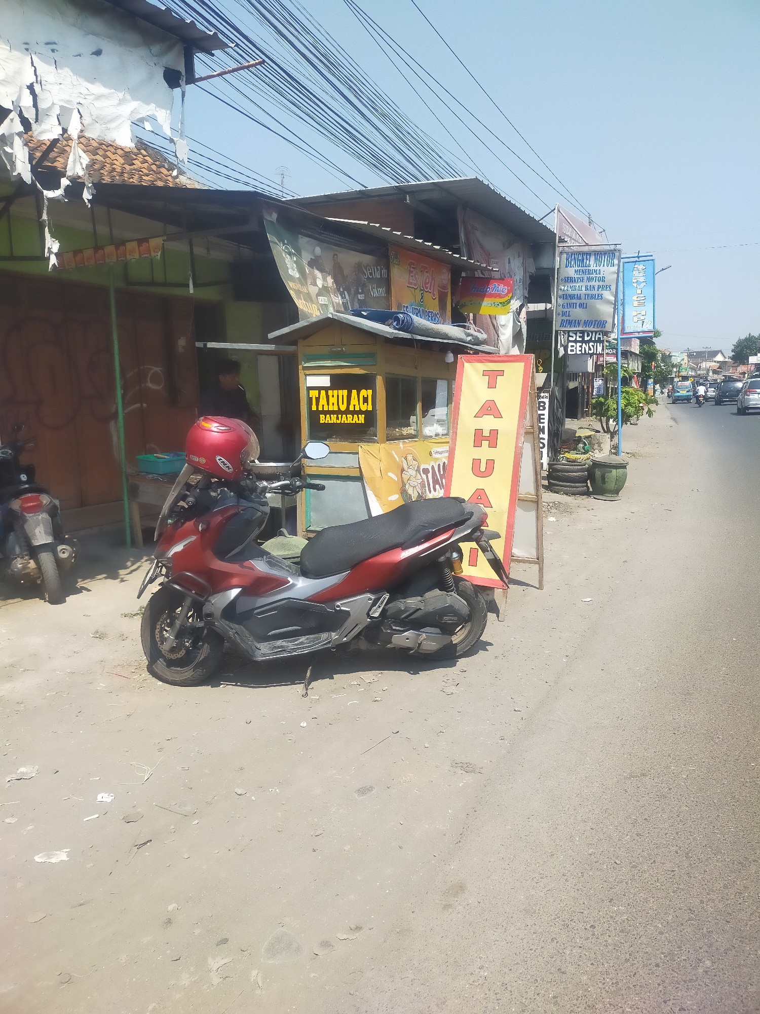 The image depicts a street scene in Talang, Indonesia, featuring a small food stall selling "Tahu Aci Banjarari". A red and silver scooter with a red helmet is parked in front of the stall. In the background, a motorcycle repair shop ("Bengkel Motor") and a convenience store ("Rima Motor Sedia Bensin") are visible. The street is lined with various shops and buildings, and several vehicles, including cars and motorcycles, are on the road. The sky is clear and blue, indicating a sunny day. The overall environment appears to be an urban or semi-urban setting with typical street commerce and traffic.