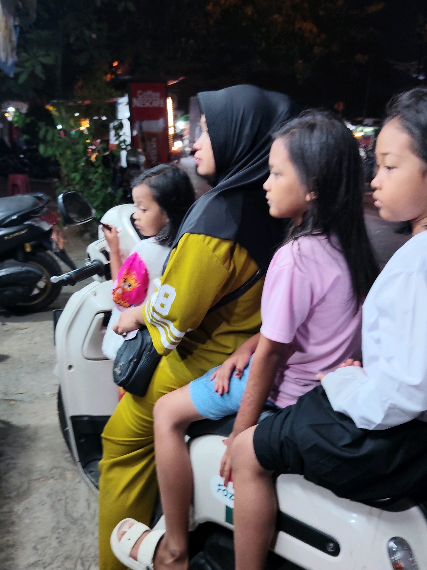 In Purwakarta, Indonesia, four individuals, a woman and three children, are depicted on a white scooter at night. The woman, wearing a black hijab and a mustard-yellow outfit with white stripes, is seated in the middle of the scooter. To her left, a young girl in a pink shirt with a character print is looking forward. To her right, two children are seated. The child closest to the woman is wearing a pink t-shirt and blue shorts, and the child furthest to the right is wearing a white shirt and black shorts. The scooter is parked on a paved surface, with a motorcycle visible in the background to the left. The setting appears to be a street or roadside, with trees and illuminated signs suggesting an urban environment. The lighting is dim, with artificial lights from the signs and possibly streetlights creating a nighttime ambiance.