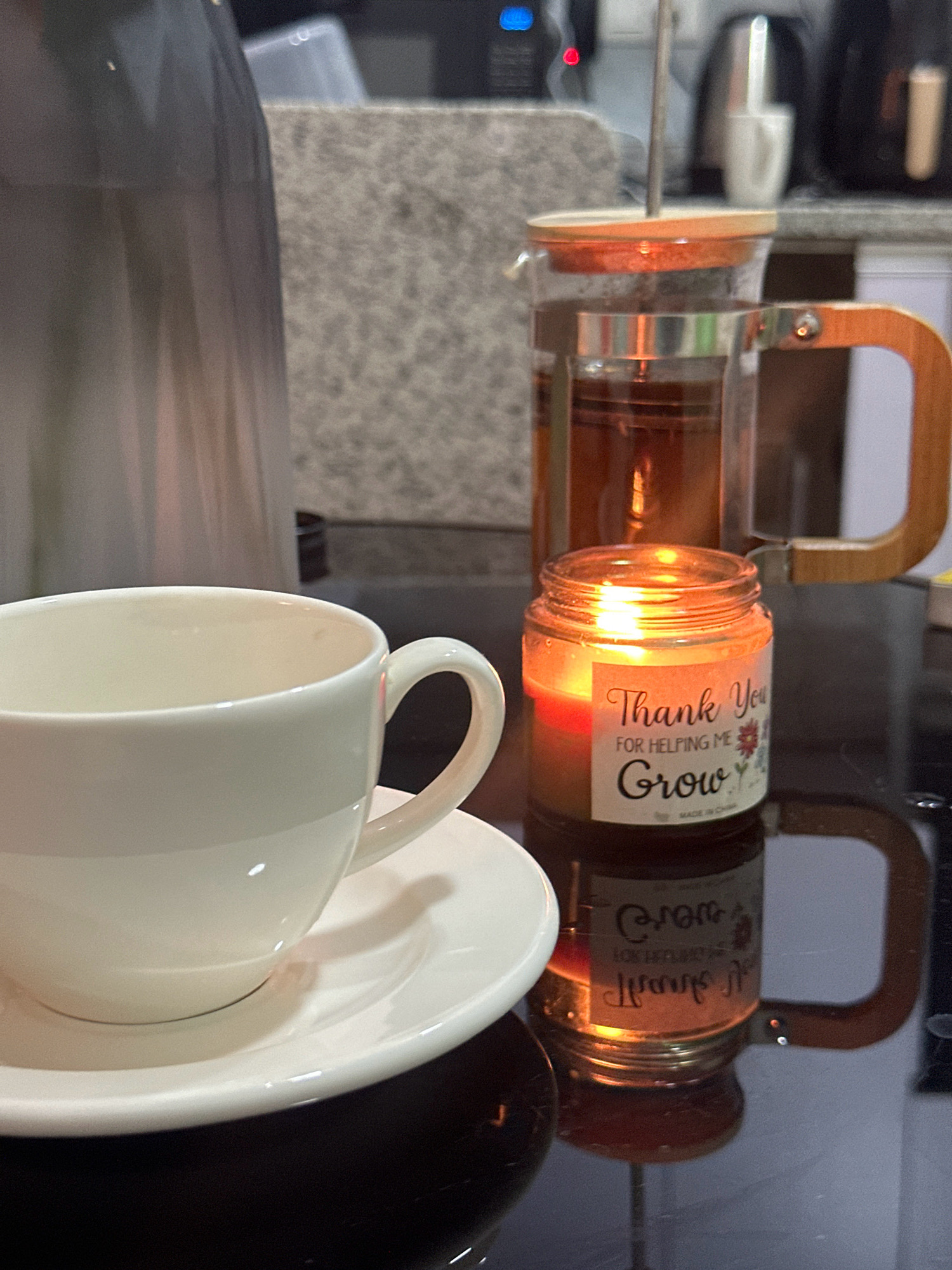The image depicts a close-up view of a domestic scene, likely a kitchen counter, with no identifiable landmarks or context to place it specifically in Lalitpur, Nepal. In the foreground, a white ceramic teacup and saucer are positioned on a dark, reflective surface. To the right of the teacup is a small glass jar candle that is lit, casting a warm glow. The label on the candle reads "Thank You For Helping Me Grow". Behind the candle and teacup, a glass French press filled with amber-colored liquid, presumably tea, is visible. The French press has a wooden handle and lid. Further in the background, out of focus, are kitchen appliances and a blurred gray textured surface. The overall atmosphere is calm and cozy, suggesting a moment of relaxation.