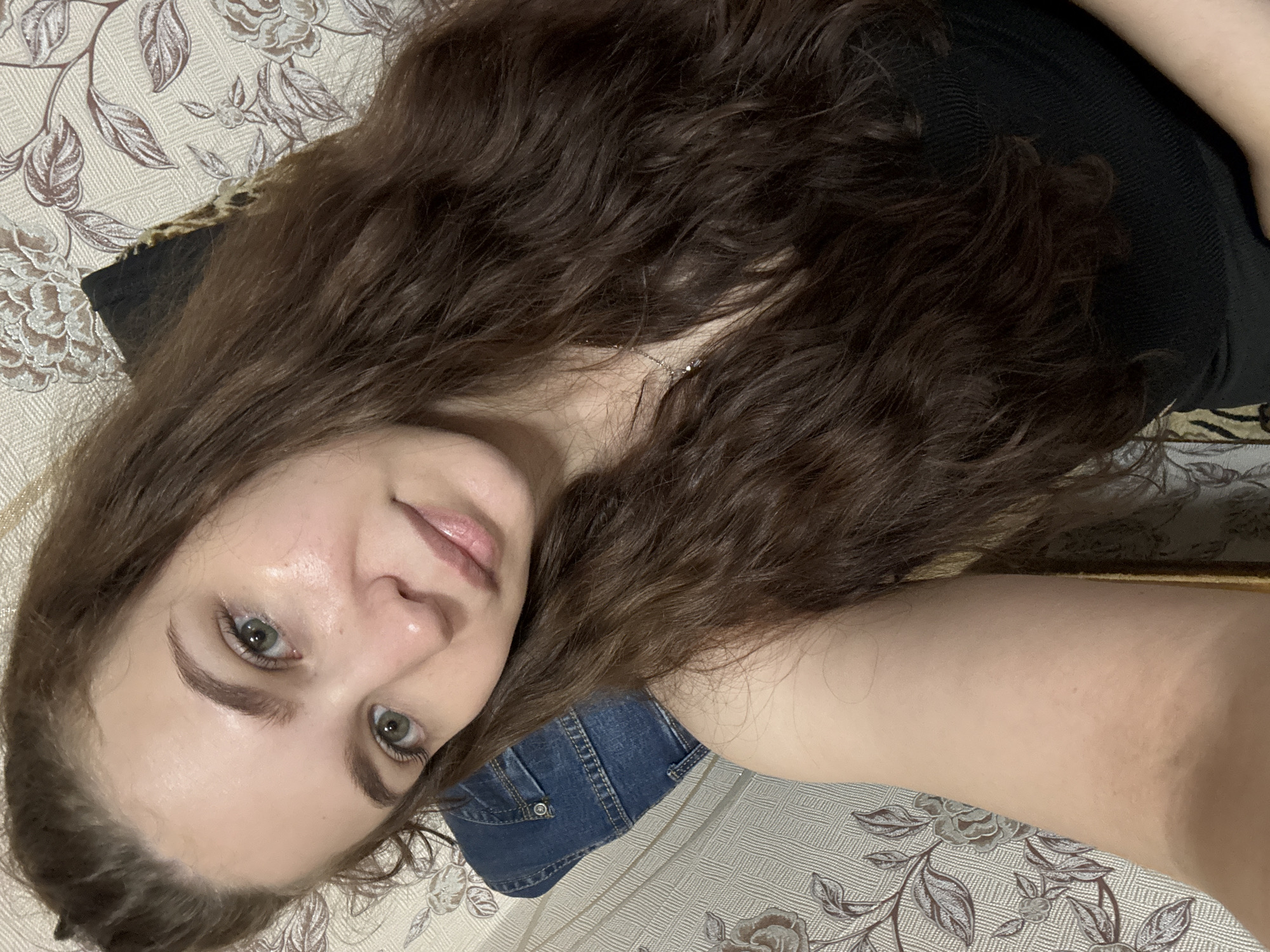 A woman with long brown curly hair is lying on a patterned surface. She is wearing a black top and denim jeans. Her face is visible, and she has green eyes and a light complexion. The background features a floral patterned surface. The location is not discernible from the image.
