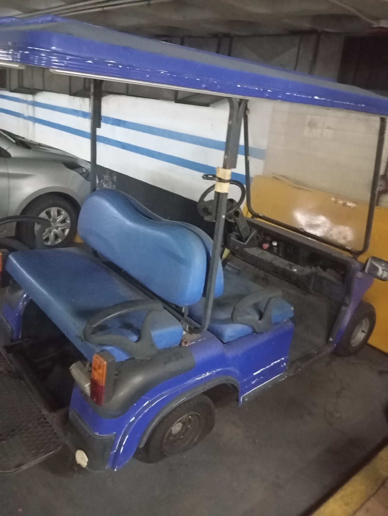 The image depicts a blue golf cart parked in what appears to be an indoor parking garage. The cart has a blue canopy and blue seats. A silver car is parked behind the golf cart. The garage has a concrete ceiling, and the wall is painted with horizontal blue and white stripes. The scene is likely an indoor parking facility. It's impossible to determine if this is located in Buenos Aires, Argentina, without additional information or context clues.