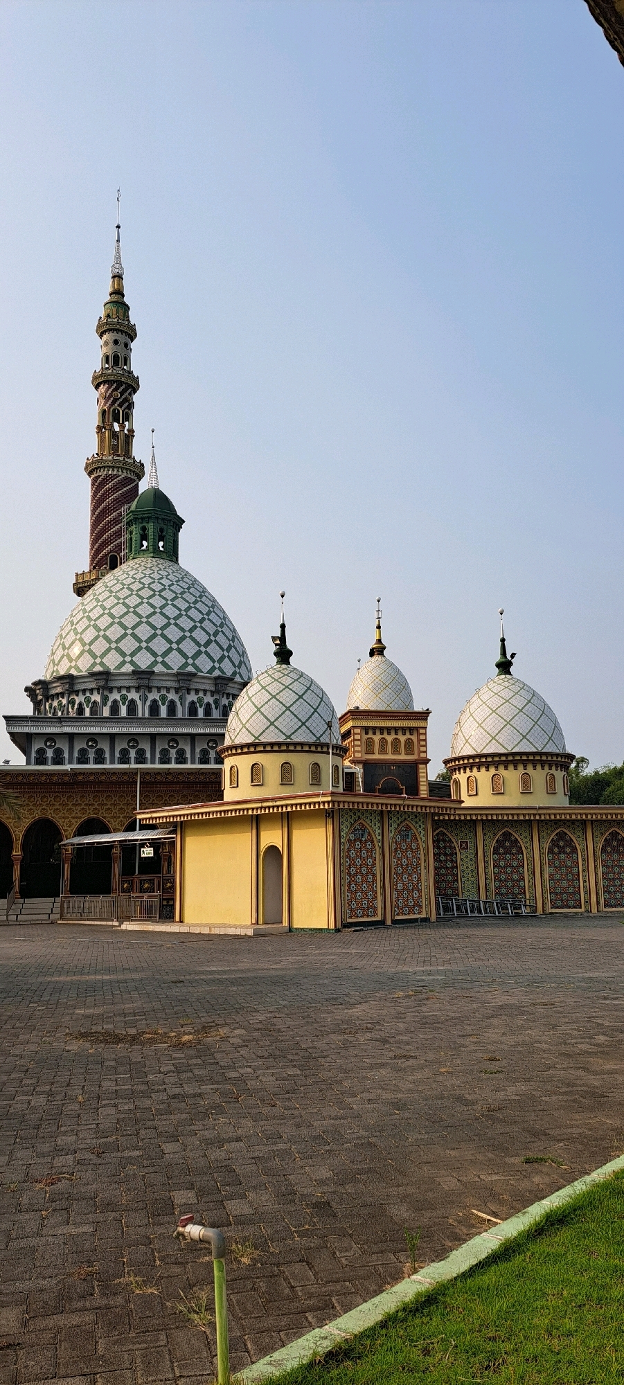 The image displays a mosque with a tall, slender minaret and multiple domes of varying sizes. The main dome is large and green with white diamond patterns, while smaller domes are white with green patterns. The building features arched windows and doorways, and is decorated with colorful tile work. In the foreground, a paved courtyard leads to the mosque, with a patch of green grass and a water faucet visible on the left. The sky is clear and blue, suggesting daytime. The mosque is located in the city of Baujeng, Indonesia. There are no people visible in the image.