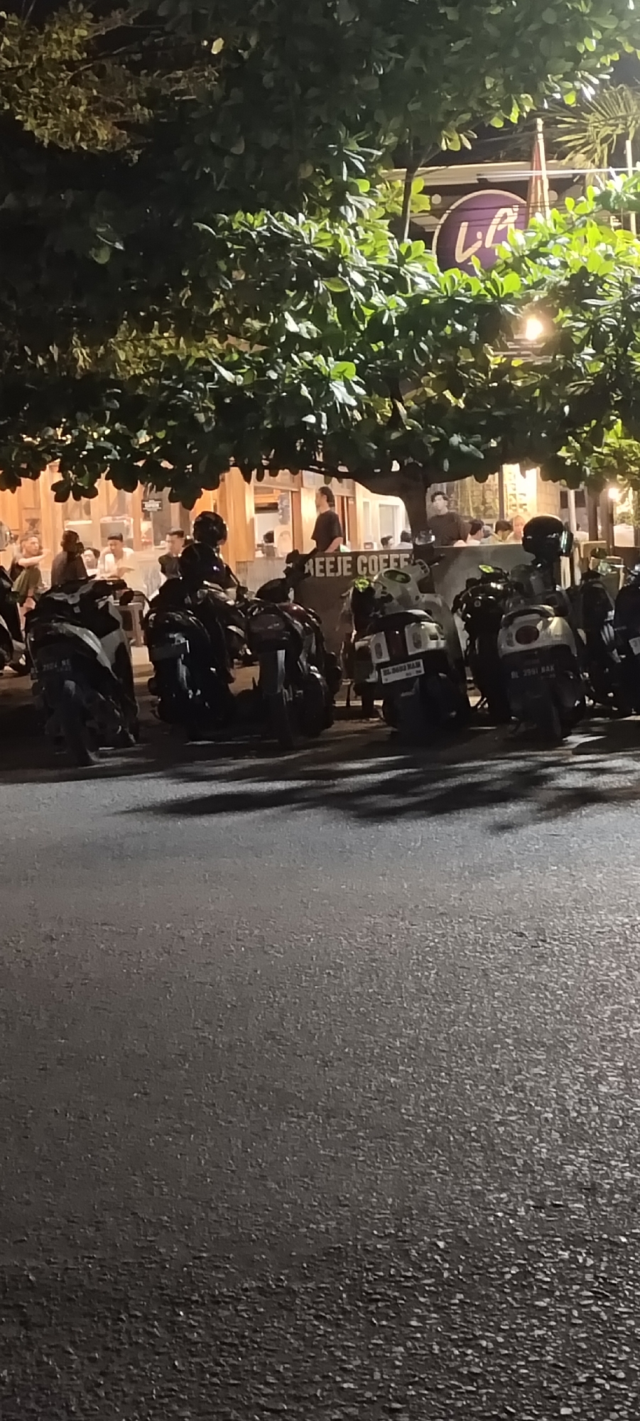 The image shows a street scene at night in Lhokseumawe, Indonesia. Parked scooters line the edge of the road in front of what appears to be a coffee shop, identified by a sign that reads "REEJE COFFEE." A sign with the letters "LA" in a stylized design is visible in the upper right, above the tree line. People are sitting at tables inside the coffee shop, and some are standing near the scooters. The road surface is asphalt. The lighting is mixed, with street lights and illumination from the coffee shop.