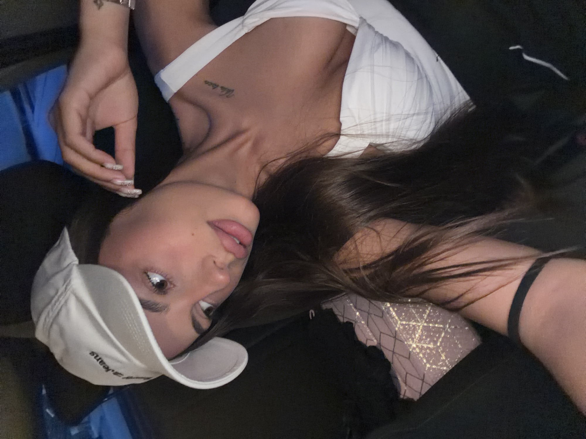 A young woman with dark hair, wearing a white baseball cap and a white top, is captured in a close-up selfie. She is positioned in a vehicle, with the interior of a car visible in the background. The woman has her hand raised to her head, with manicured fingernails adorned with rhinestones. A tattoo is visible on her collarbone. The lighting suggests it is either evening or nighttime. The image does not contain any information to suggest the location is the city of Karnobat, Bulgaria.