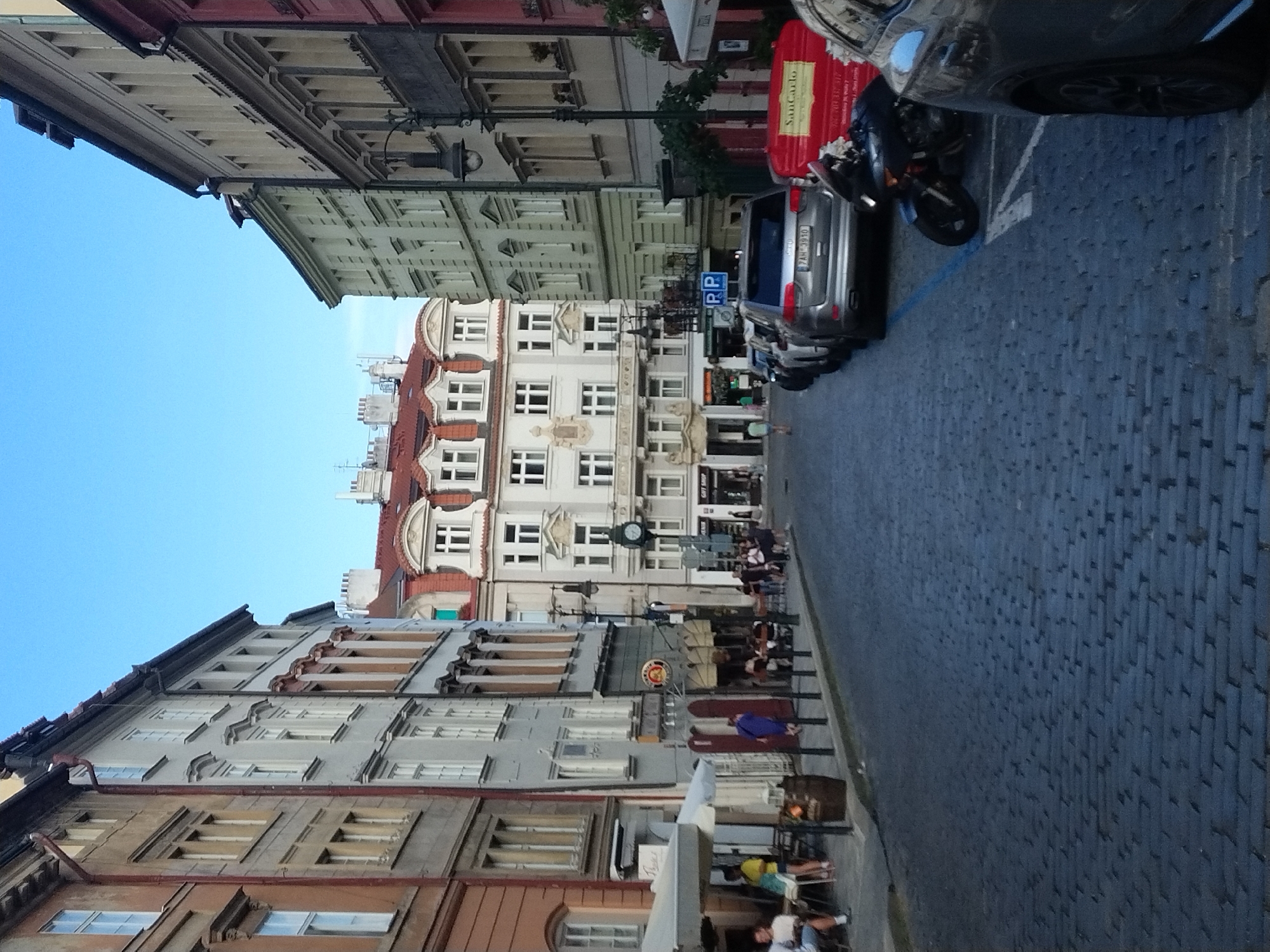 The image depicts a cobblestone street in Prague, Czechia, with a clear blue sky overhead. On the left side of the street, there are buildings with multiple stories and windows, some of which appear to be shops or cafes with outdoor seating. A white umbrella covers some tables. People are seated at tables and walking on the sidewalk. On the right side of the street, there are more buildings, a lamppost, and parked cars. A motorcycle is parked in a designated spot. The buildings showcase architectural styles common in older European cities, with facades of varying colors and decorative elements. The street is paved with dark cobblestones.