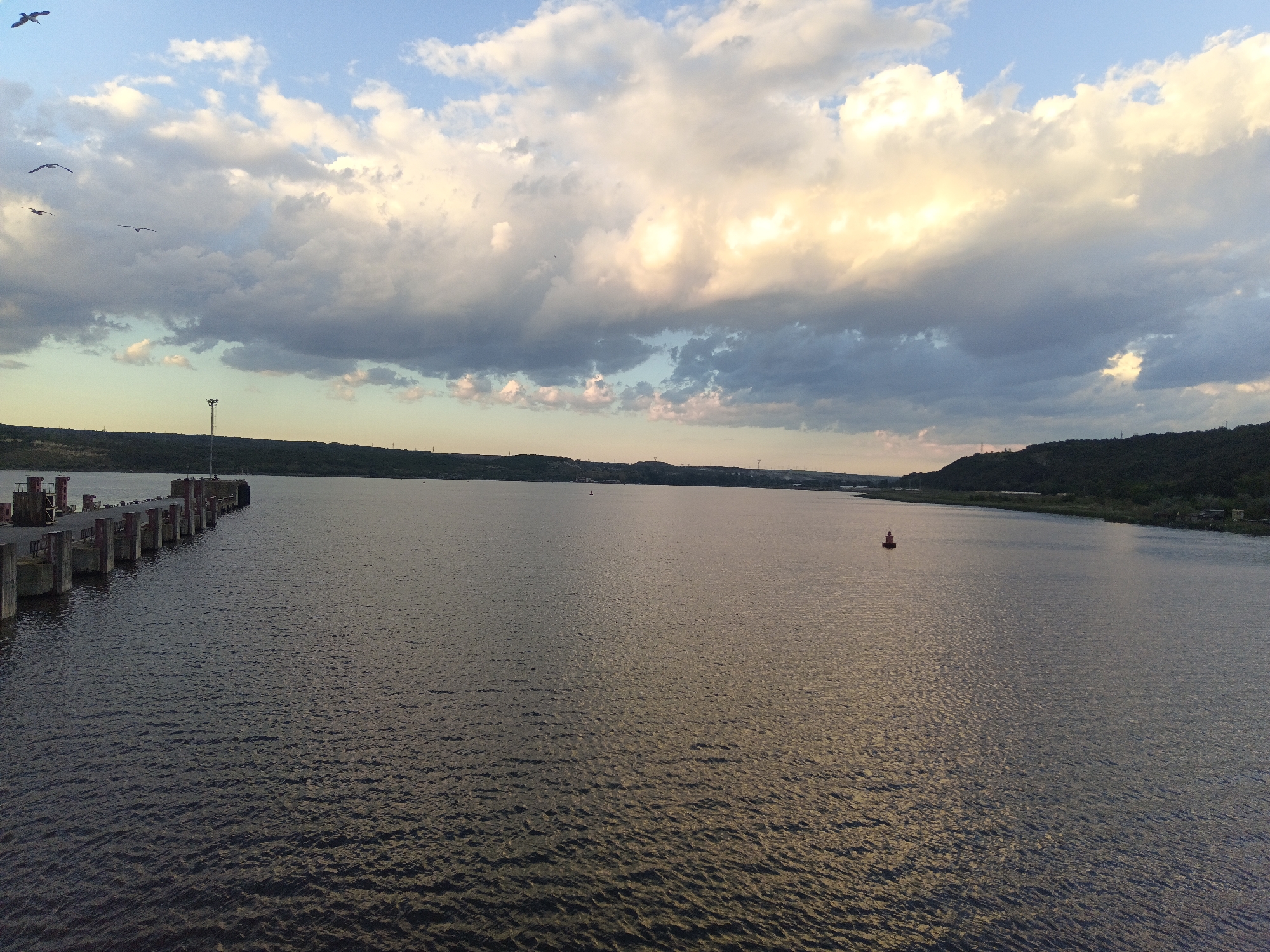 The image shows a wide body of water, likely the Beloslav Lake in Bulgaria, based on similar landscape features. A pier or dock extends from the left edge of the frame into the water. A single red buoy is visible in the middle of the water. The shoreline in the distance features a hilly landscape with sparse vegetation. The sky is filled with clouds, ranging from light and bright to darker and more ominous. There are some birds flying in the sky on the left.
