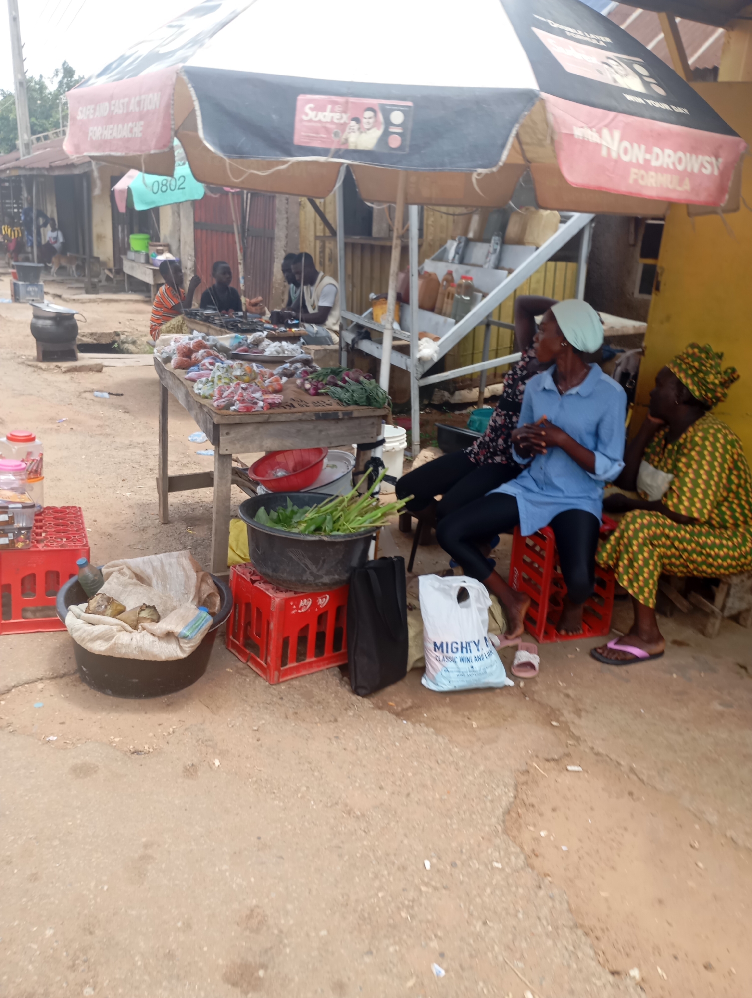 This is a full, eye-level, medium shot of an outdoor market stall in Shendam, Nigeria. A large, black and red umbrella with a Sudrex advertisement is positioned over the stall. Several people are present, including two women and at least three men. Some are seated on crates, while others stand or are partially visible behind the stall.
The stall itself features a wooden table laden with various items, including what appear to be wrapped food products, onions, and leafy greens. To the left of the table, a large black basin filled with leafy greens rests on a red crate. Another large, dark basin, partially covered with a woven sack, is situated to the left, containing some produce. Various plastic containers and red crates are scattered around the scene. The ground is made of dirt and sand. The overall atmosphere suggests a busy marketplace.