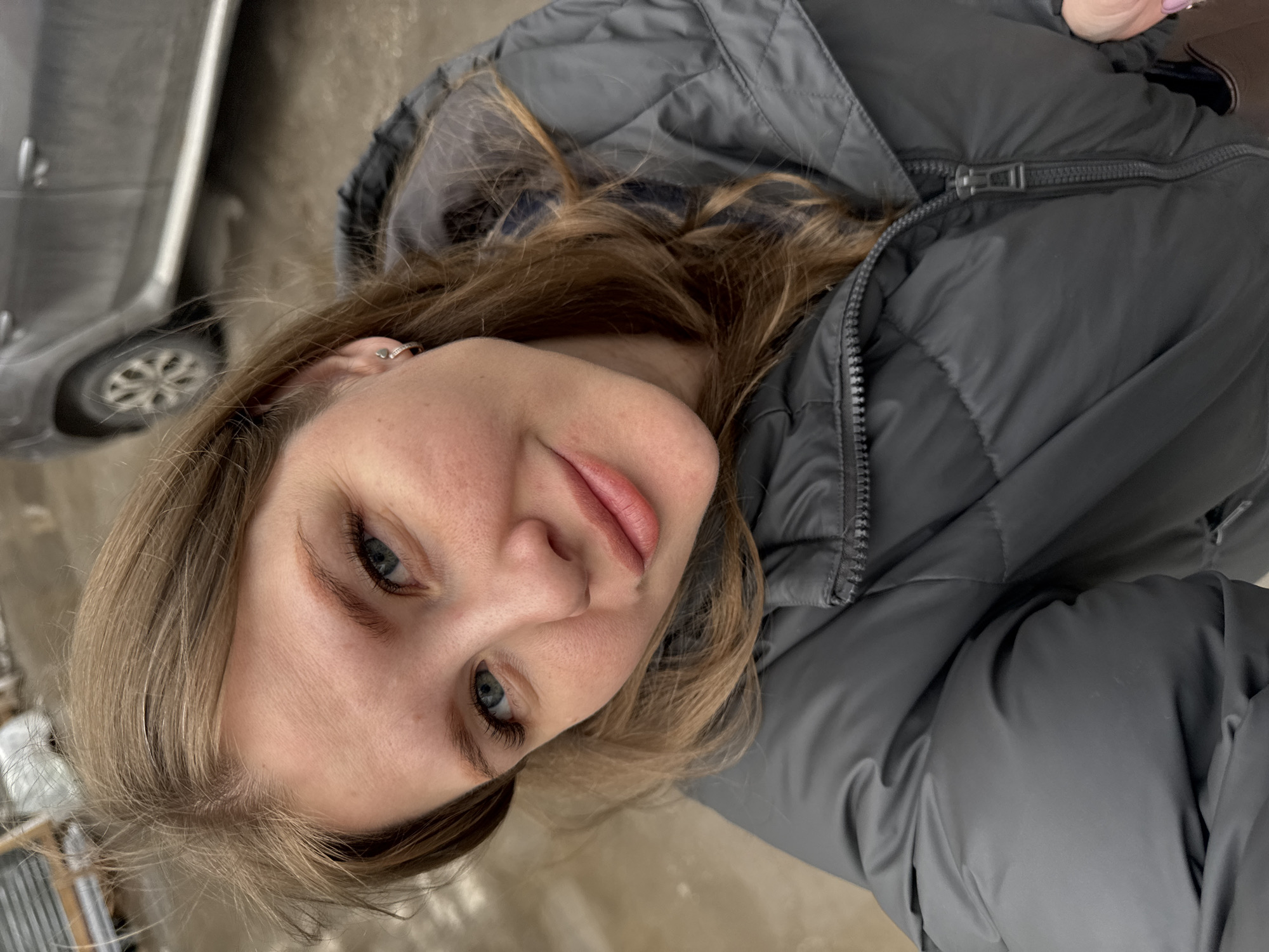 A close-up, eye-level shot captures a young woman outdoors, likely in Perm, Russia, given the context. The image is angled, showing her face looking slightly upwards and to the side, directly at the camera, with a calm and gentle expression. She has light brown to blonde hair, styled loosely around her face, and striking blue eyes. Her lips are lightly colored with a pink hue.
She is wearing a practical grey puffer jacket, suggesting cooler weather. A small, silver-colored heart-shaped earring is visible in her left ear. The jacket's zipper and quilted texture are evident.
In the blurred background, partially visible to the left, is a grey car. A wheel and a portion of the car's side are discernible, indicating an urban or semi-urban outdoor setting like a street or parking area. The ground surface is indistinct but appears a light brown or grey, possibly pavement or damp earth. The lighting is natural and soft, characteristic of daytime. There is no visible text in the image.