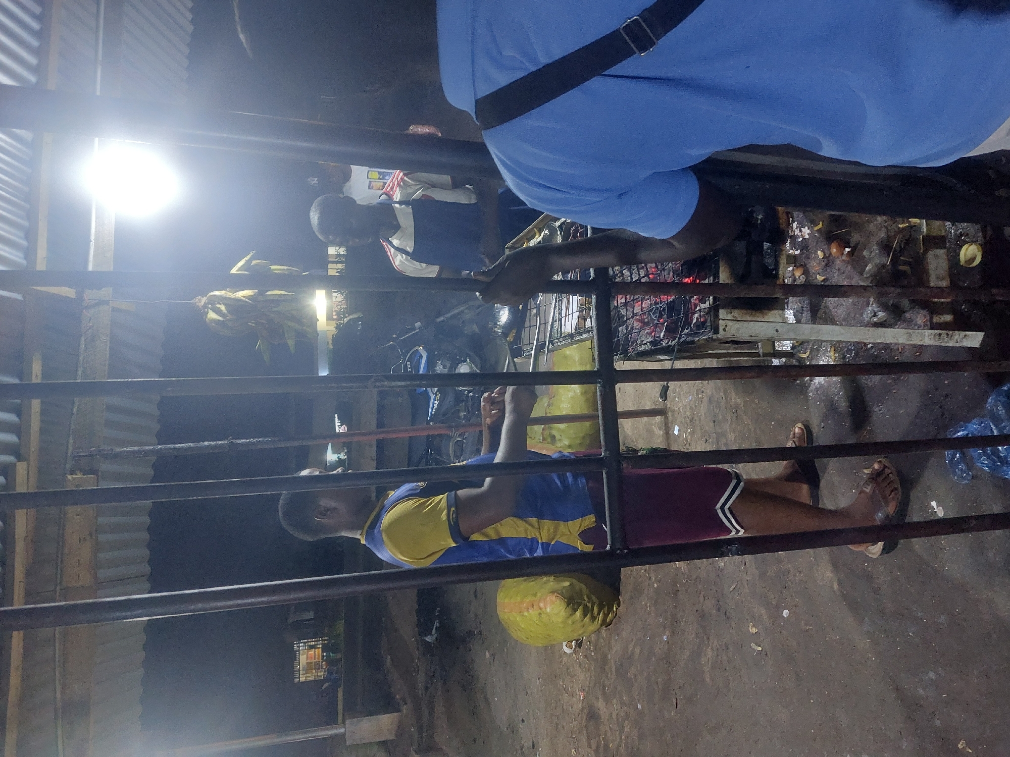 The scene captured in the image is an outdoor area at night in Dar es-Salaam, Tanzania. The environment is dimly lit by a bright light in the background. A metal structure of horizontal bars is in the foreground. A man wearing a blue and yellow shirt and maroon shorts is lying down with a yellow sack next to him. Another man, wearing a blue shirt with a black strap across his shoulder, is standing near a barbecue. A third man is also standing near the barbecue. They appear to be grilling food over a charcoal fire. There are various objects on the grill.