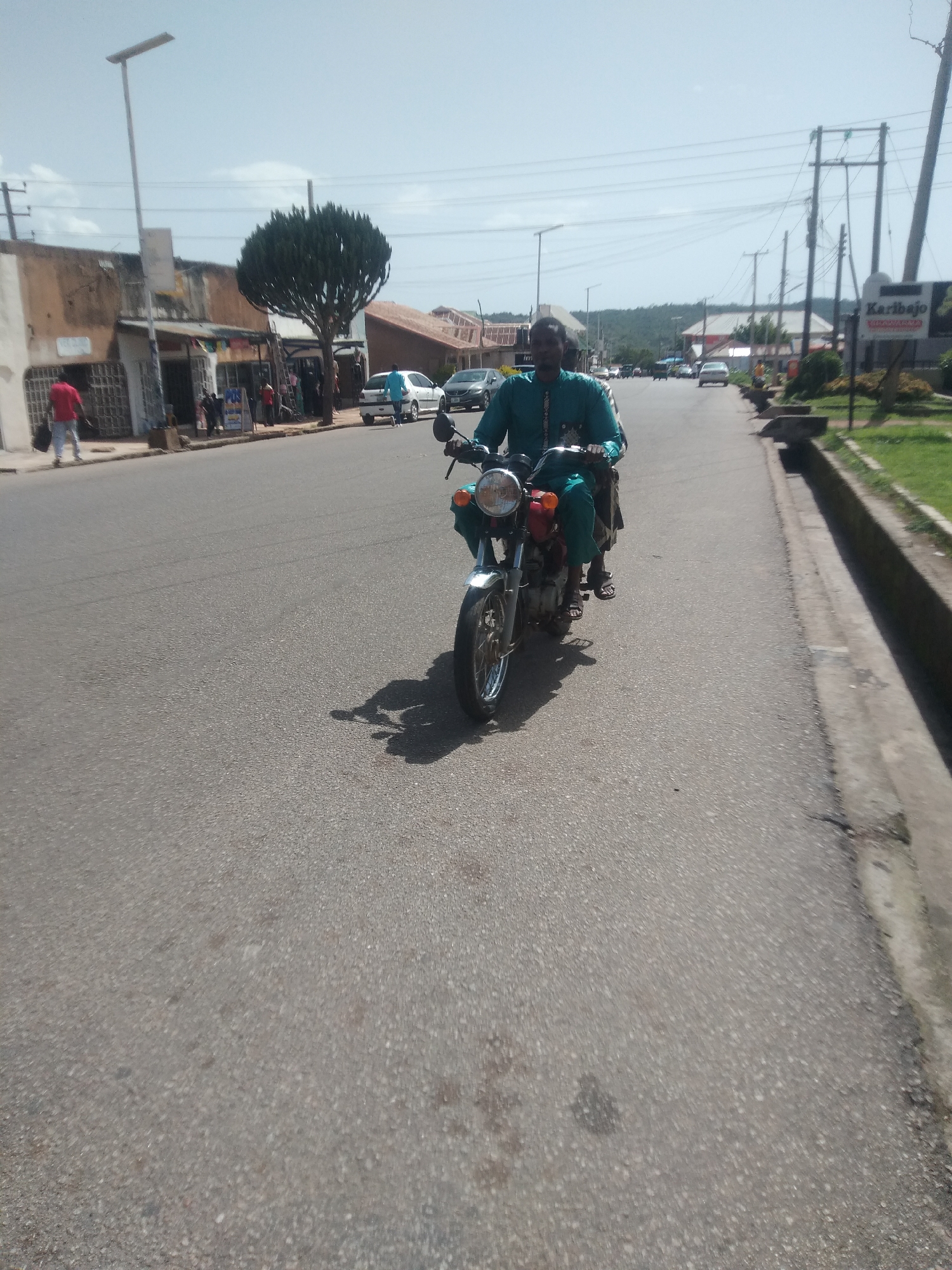 A man is riding a motorcycle down a paved road in Jos, Nigeria. He is wearing a teal colored traditional outfit. The motorcycle is red and silver with chrome accents. The man is looking straight ahead. The scene appears to be a commercial area with buildings and shops lining the left side of the road. A tall, thin evergreen tree stands in front of some of the shops. Power lines are visible overhead. Several cars and other motorcycles are on the road in the distance. The weather appears to be clear and sunny, suggesting it is daytime. A sign with the text "Karibajo" and "BAKARO" is visible on the right side of the road. The overall mood seems to be ordinary daily life in a busy town.