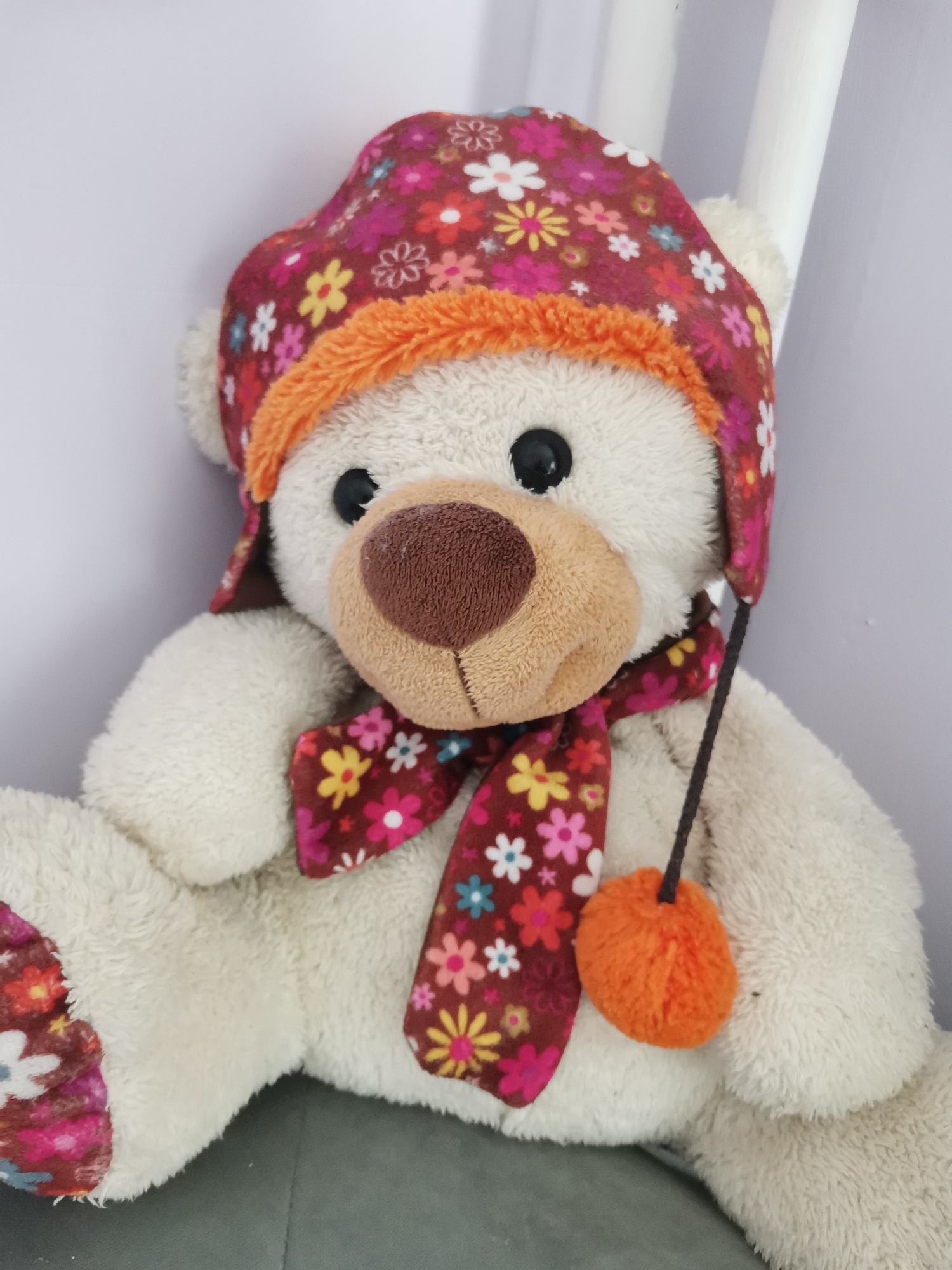A cream-colored plush teddy bear is depicted in a seated, upright position. The bear features black button eyes and a light brown, velvet-textured nose and snout. It wears a deep maroon hat adorned with a vibrant floral pattern in white, yellow, pink, orange, and light blue, accented with an orange fluffy trim around the face opening. A matching floral scarf is tied around its neck. A dark brown string with a bright orange fluffy pom-pom hangs from the right side of the hat. The bear's left foot is partially visible, showing a patch with the same floral pattern. The background consists of a light off-white or light gray wall, with two narrow, vertical white cylindrical objects partially visible on the right side. The bear rests on a dark gray, possibly upholstered, surface. The image provides no discernible visual information to determine a specific geographical location, such as Sofia, Bulgaria.