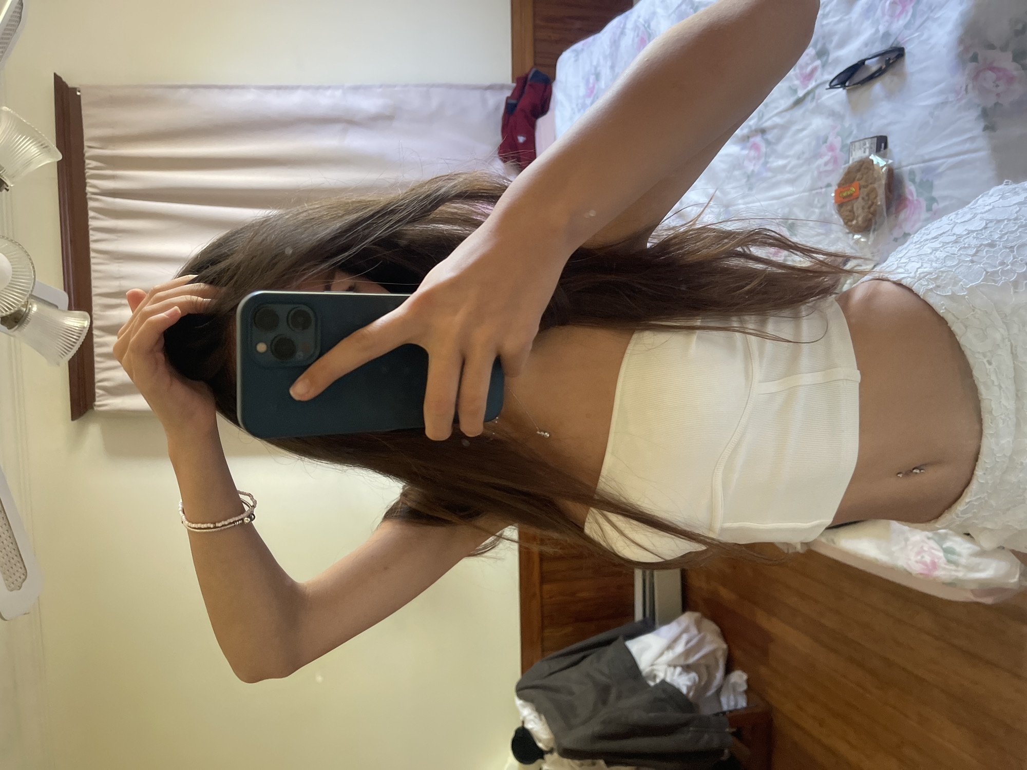 The image shows a young woman taking a mirror selfie in what appears to be a bedroom. She is wearing a white, strapless top and a white lace skirt, and has her hair down. She is holding a smartphone in her right hand, partially obscuring her face. A bracelet is visible on her left wrist. In the background, a bed with a floral-patterned comforter is visible, as are a closet with clothing and a dresser. The room has wooden floors and light-colored curtains. The location is not specified in the image itself.
