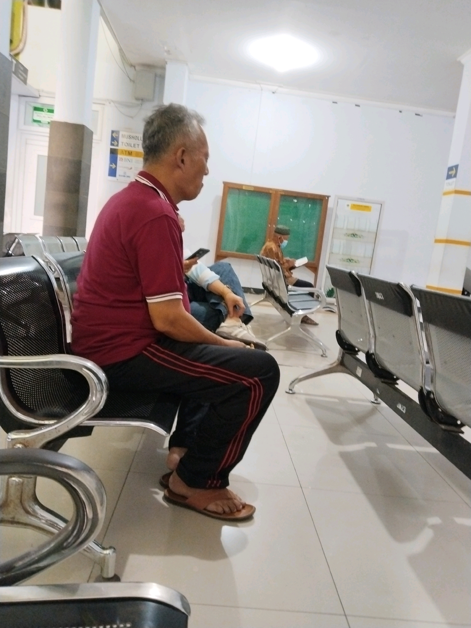 The image depicts a waiting area, likely within a public building such as a hospital or government office, in Trenggalek, Indonesia. In the foreground, an elderly man of East Asian descent is seated on a metallic chair. He has gray hair and is wearing a maroon t-shirt with a white collar and stripe accents, and dark pants with red stripes down the sides. He is wearing brown sandals and is looking to his right. Behind him, other individuals are seated, and rows of metallic chairs are visible, some with black mesh seats. A sign on a pillar indicates directions to "MUSHOLLA TOILET" and "ATM BNI". In the background, a green board is mounted on the wall, and a door with a yellow sign is visible. The floor is tiled in a light color. The lighting suggests an indoor environment with overhead illumination.