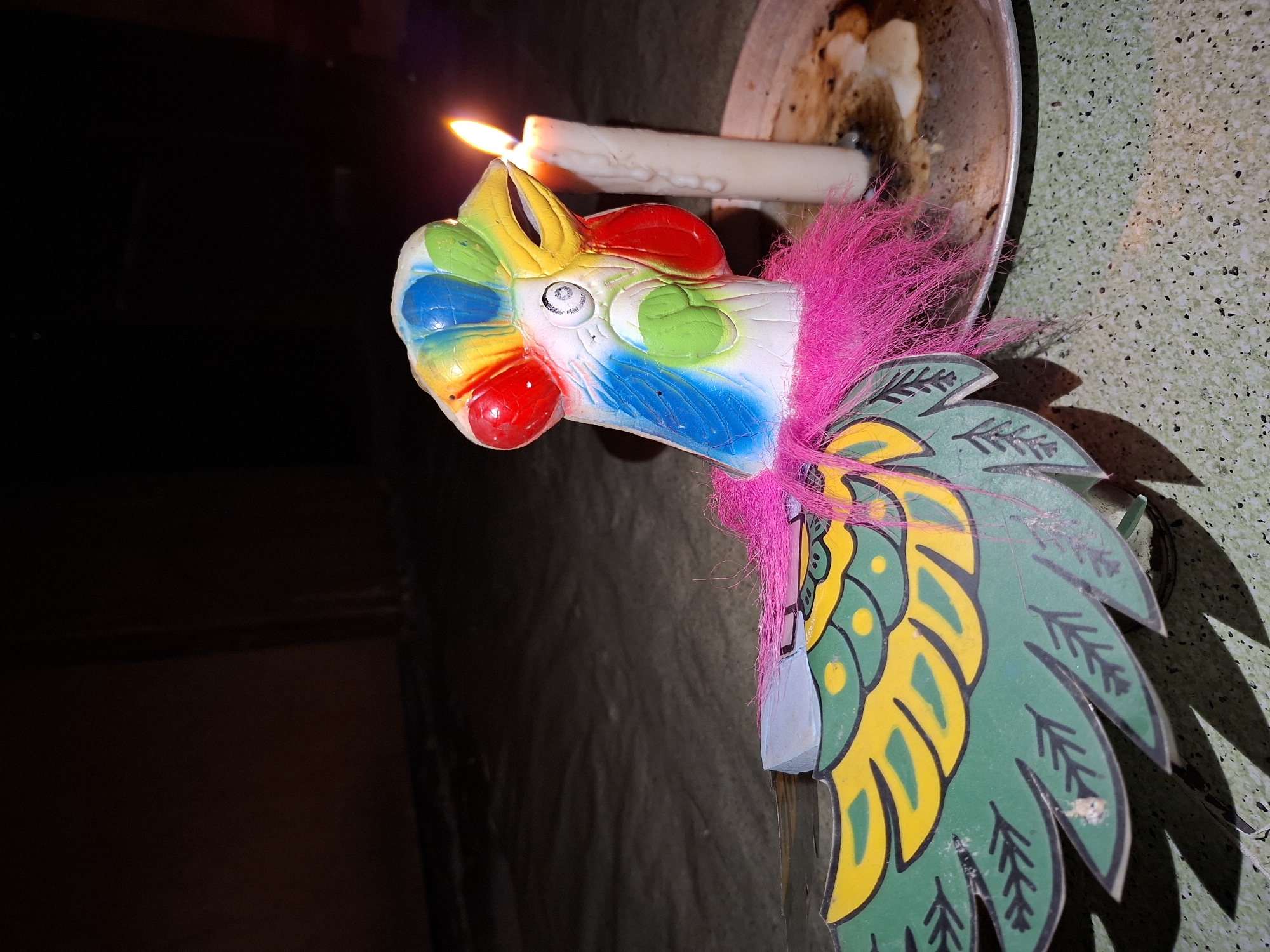 A lit candle sits in a metal container, with its flame illuminating a colorful, stylized rooster figurine. The figurine has a red beak, a blue neck, a white body with green and yellow circles, a rainbow-colored head crest, and pink feathery accents. The back of the rooster is adorned with green and yellow feathers that have black arrow-like markings. The background is dimly lit. Based on the reference image, the location is unspecified, but the prompt mentions Lhokseumawe, Indonesia. Therefore, the scene is presumed to be located in Lhokseumawe, Indonesia.
