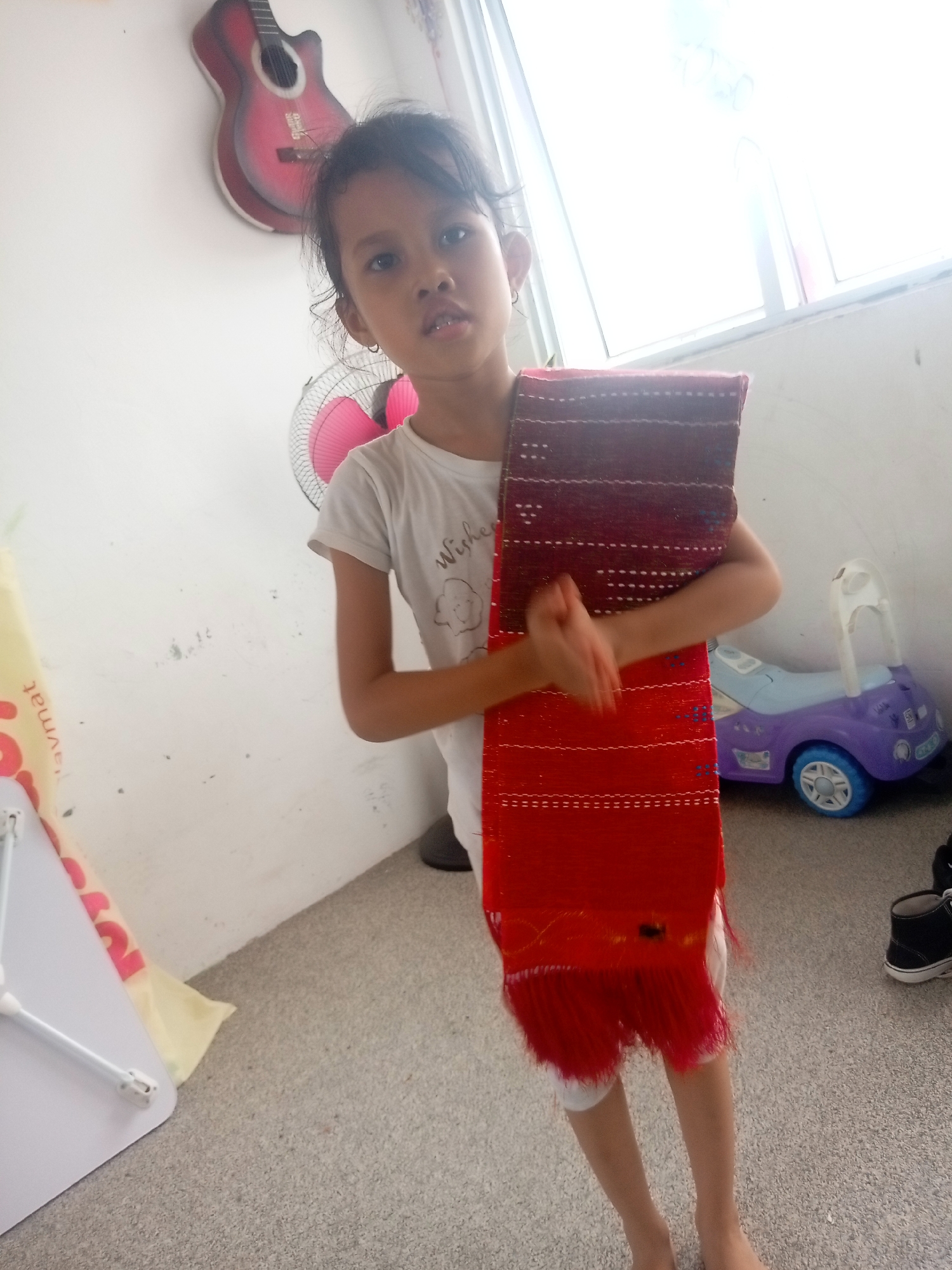 A young girl stands in a room, holding a woven fabric with red and maroon colors and fringe. She is wearing a white t-shirt with a graphic print and white leggings. The room has a light-colored carpeted floor and white walls. In the background, there is a pink fan, a red guitar mounted on the wall, a small purple toy car, and a white laptop table. Sunlight streams in from a window. This scene is likely in Batam, Indonesia, given the context.