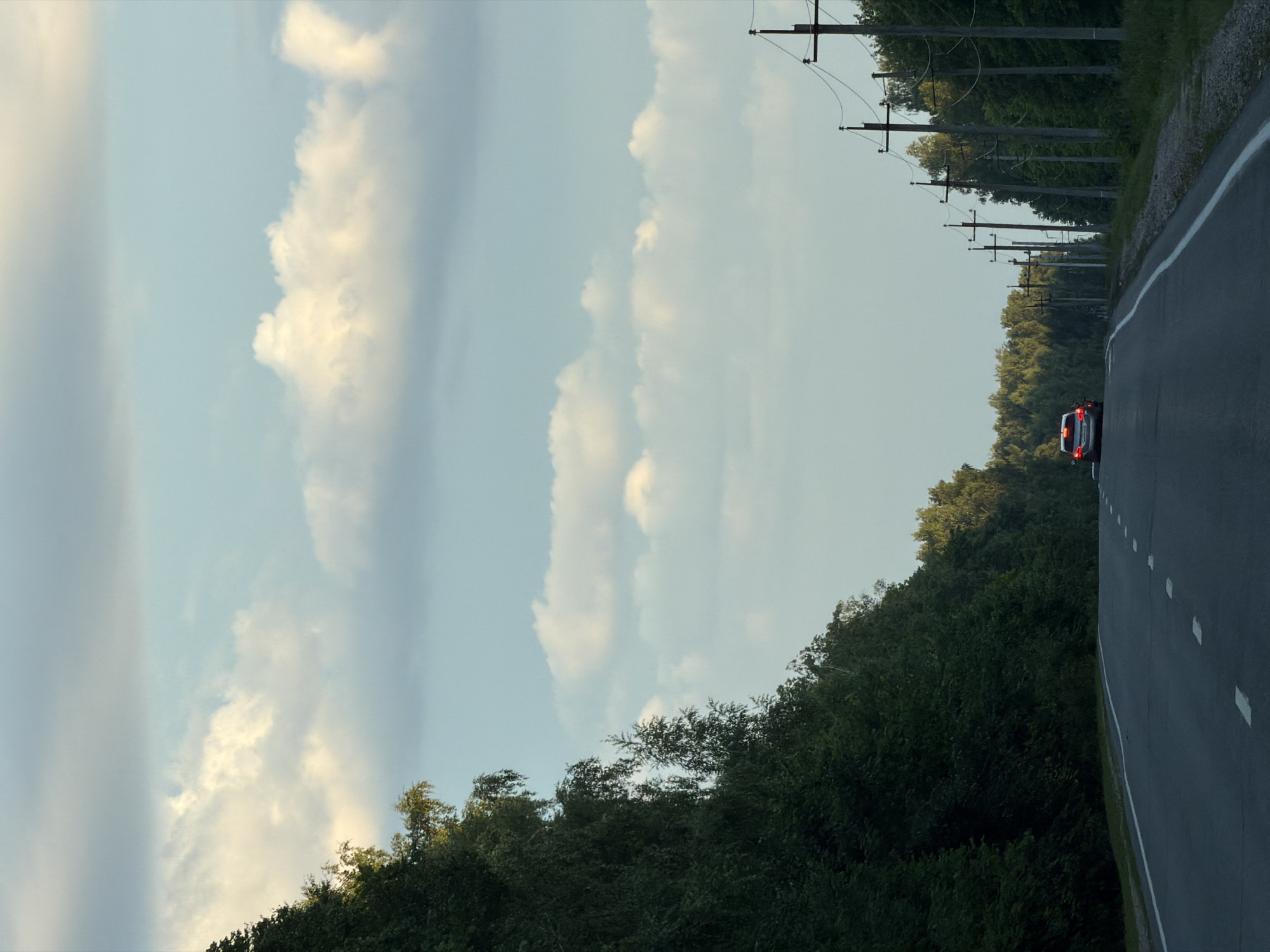 A silver sedan with its brake lights illuminated is driving on a two-lane asphalt road. The road is bordered on the left by a dense, dark green forest and on the right by a line of wooden utility poles and a further forest. The sky is light blue with scattered, fluffy white clouds. The setting appears to be a rural or semi-rural area, possibly on the outskirts of Laishevo, Russia, given the presence of the road, forest, and utility infrastructure typical of such regions.