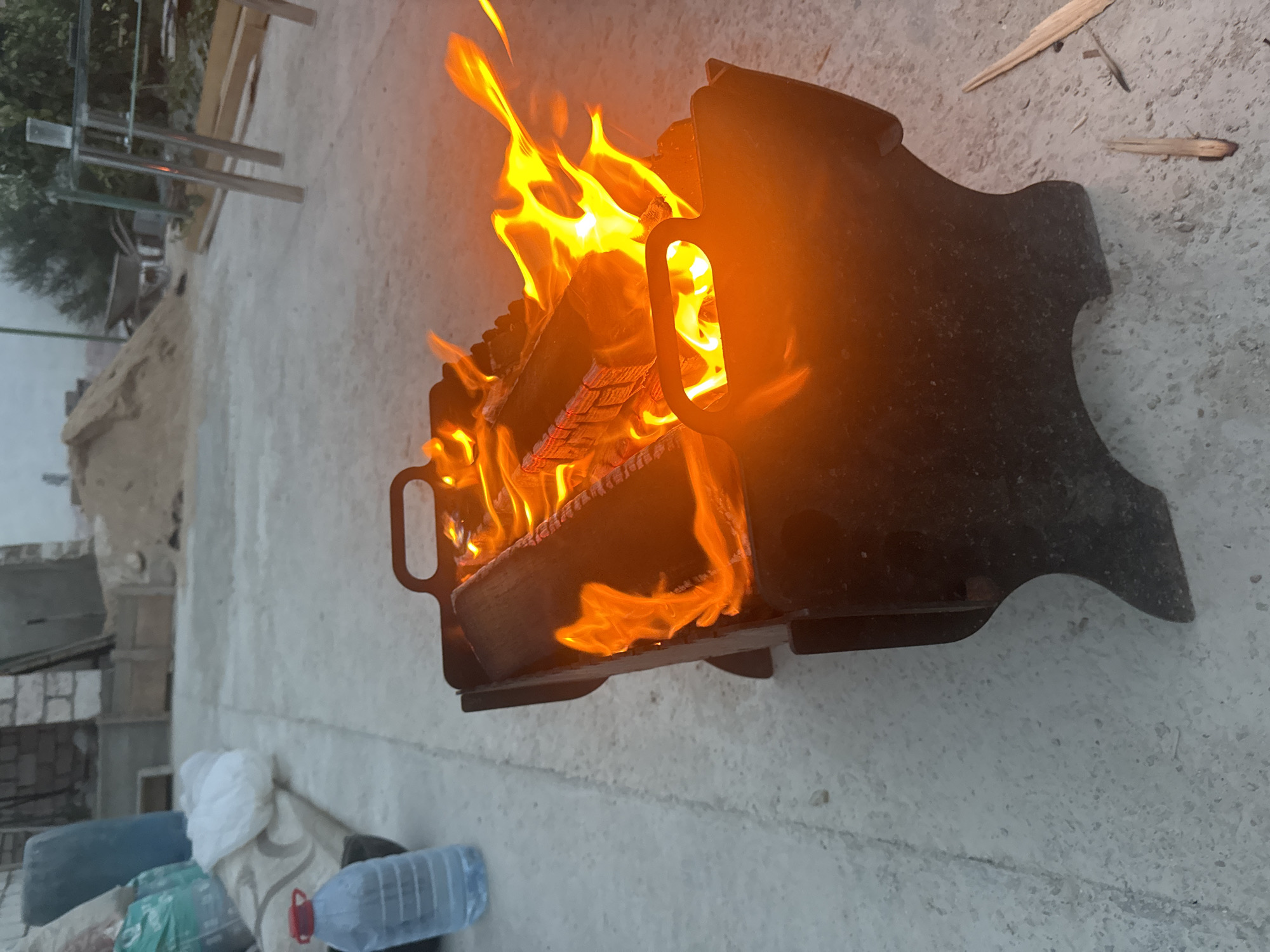 An outdoor scene in Atameken, Kazakhstan, features a portable black metal fire pit actively burning logs. Flames, bright orange and yellow, rise from the fire pit, illuminating the surrounding area. To the left of the fire pit, a large plastic water bottle with a red cap is visible. In the background, construction materials including sand, concrete blocks, and lumber are present. The ground is paved with a light-colored concrete surface. The lighting suggests it is either dawn or dusk.