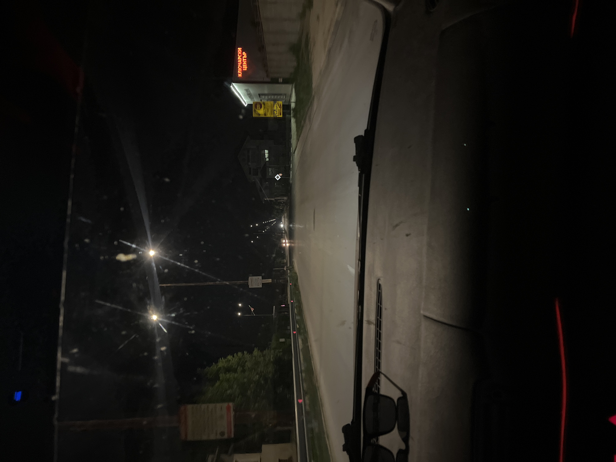 The image shows a night-time view from inside a vehicle, looking out onto a street in Yasen, Bulgaria. The perspective is from the driver's seat, with the windshield and dashboard visible. The road ahead is illuminated by streetlights and the headlights of other vehicles. Trees and buildings line the street, and a few signs are visible. The overall mood is somewhat eerie due to the darkness and the low quality of the image.
One prominent object in the scene is a pair of sunglasses resting on the dashboard. On the left side of the image, a building with a bright red neon sign is visible. The sign displays text in Cyrillic script, which appears to read "КЛЮЧАРСКИ ЦЕНТЪР," meaning "Locksmith Center."
The time of day is clearly night, as indicated by the darkness and the artificial lighting. The weather appears to be clear, as there is no visible precipitation or fog. The scene depicts a typical street at night, with vehicles moving along the road and businesses illuminated. The absence of people suggests that it is late at night or early in the morning. The image is slightly blurry and grainy, possibly due to low light conditions or the quality of the camera used.