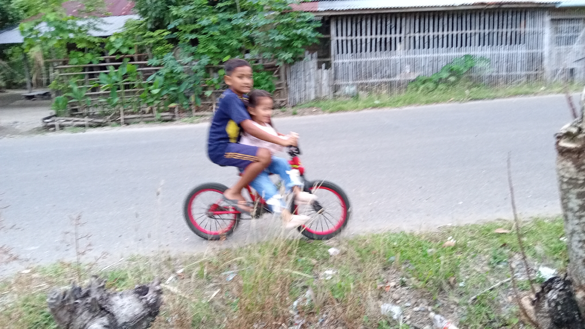 Two children, a boy and a girl, are riding a red bicycle on an asphalt road. The boy is pedaling and steering, with the girl seated behind him. Both are wearing casual clothing. The bicycle has red rims on its wheels. The scene appears to be in a rural or semi-rural area, with vegetation and a building with a corrugated metal roof visible in the background. The building's facade is constructed with vertical wooden slats. The road is bordered by grassy areas with some dried foliage and debris. This scene is located in Uteunreutoh, Indonesia.