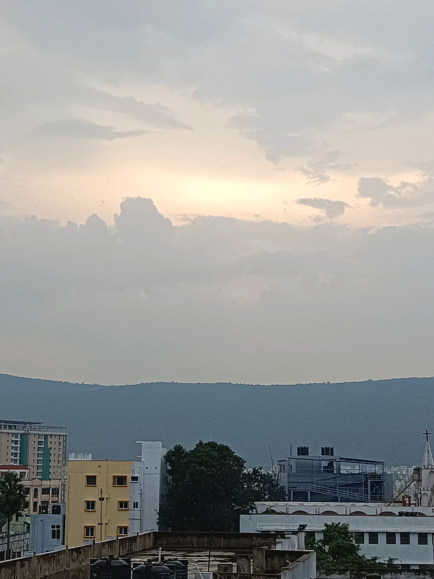 The image captures a skyline in Visakhapatnam, India, dominated by a silhouetted mountain range in the background. The sky is overcast with soft, diffused light, suggesting either dawn or dusk. In the mid-ground, a cluster of buildings of varying heights and colors form the urban landscape.
Notable structures include a tall, multi-story building with a light-colored facade and many windows on the left. Adjacent to it is a shorter, bright yellow building with a grid of windows. Further to the right, a white building with a distinctive rooftop structure that appears to be under construction, featuring scaffolding and water tanks, is visible. A white spire, possibly part of a church or religious building, topped with a cross, is also present on the right side of the frame. Several trees, including a palm tree on the left, punctuate the cityscape. The foreground shows the rooftops of lower buildings, some with water tanks. The overall impression is a typical urban view with natural topography serving as a backdrop. No people are discernible in the image.