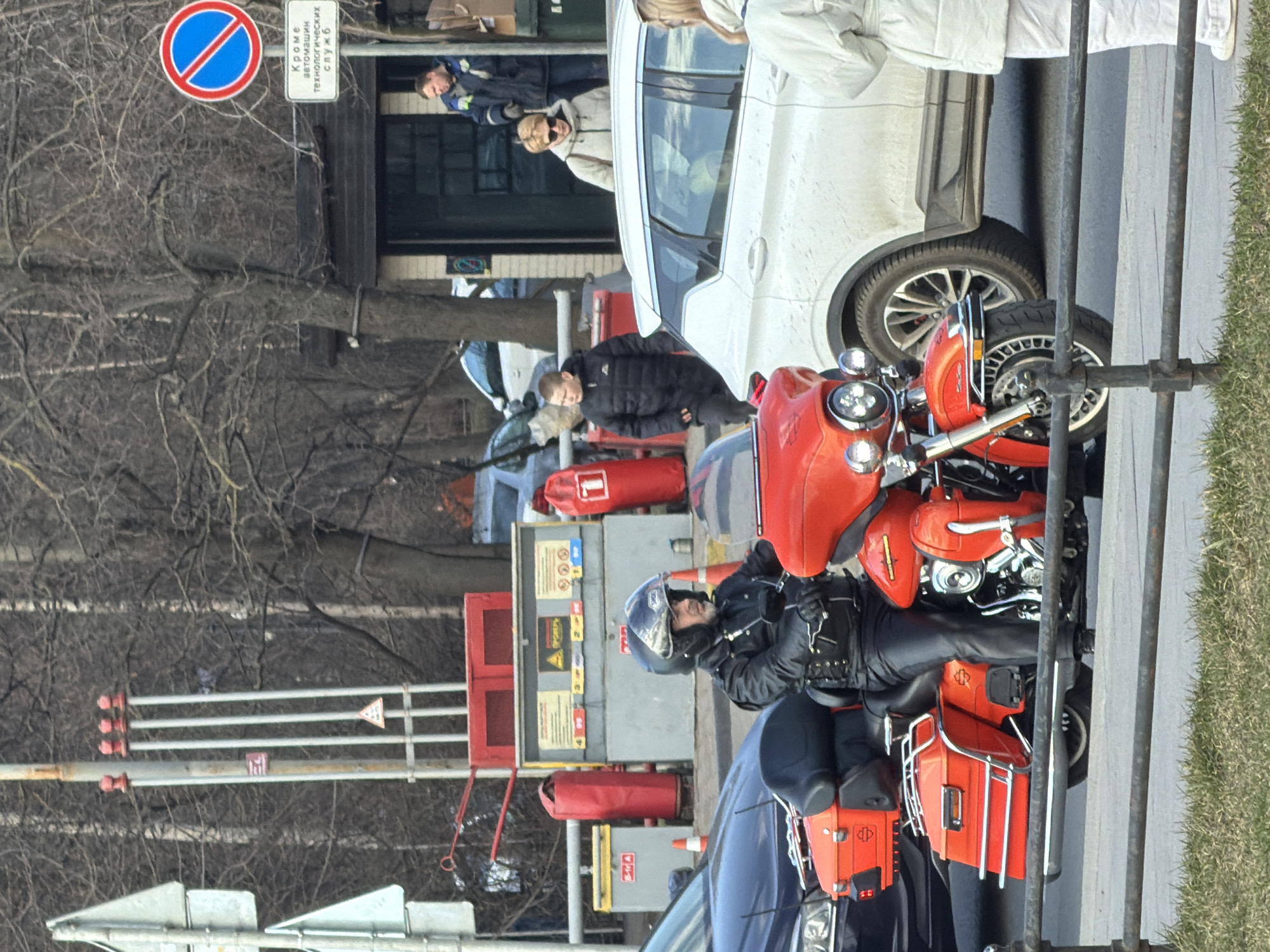 An urban street scene in Saint Petersburg, Russia, depicts a bright orange Harley-Davidson touring motorcycle stopped or stationary on asphalt pavement. A male individual, wearing a black helmet with a clear visor, a black leather jacket, black leather gloves, and black leather pants, is seated on the motorcycle. He is facing slightly right, hands on the handlebars.
In the immediate foreground, a dark metal railing separates a strip of brown-green grass from the street. To the left of the motorcycle, a dark-colored sedan is partially visible. To the right, a white SUV with multi-spoke wheels is also stopped or parked. A female individual in a light-colored padded jacket and trousers is partially visible in the far right foreground, facing away from the viewer.
Behind the white SUV, a woman with blonde hair wearing sunglasses and a light-colored padded jacket walks to the left. Further back, a young male in a black padded jacket walks towards the left. A male individual in a blue and yellow work uniform stands in the doorway of a dark building with a green door.
To the left of the scene, a utility area features red vertical bollards and gray metal boxes with yellow instructional signs, suggesting fuel pumps or a service point. Two orange traffic cones are visible near these structures. A tall pole with four vertical white pipes topped with red caps stands behind the utility area. To the far left, two gray traffic signs are partially visible.
In the mid-background to the right, a blue circular "No Entry" sign with a red diagonal bar is mounted above a white rectangular sign displaying Russian text: "КРОМЕ автомашин технологических СЛУЖБ" (EXCEPT technological service vehicles). The background is dominated by a dense arrangement of bare deciduous trees, indicating a colder season. The lighting is bright and diffuse, suggesting daytime.