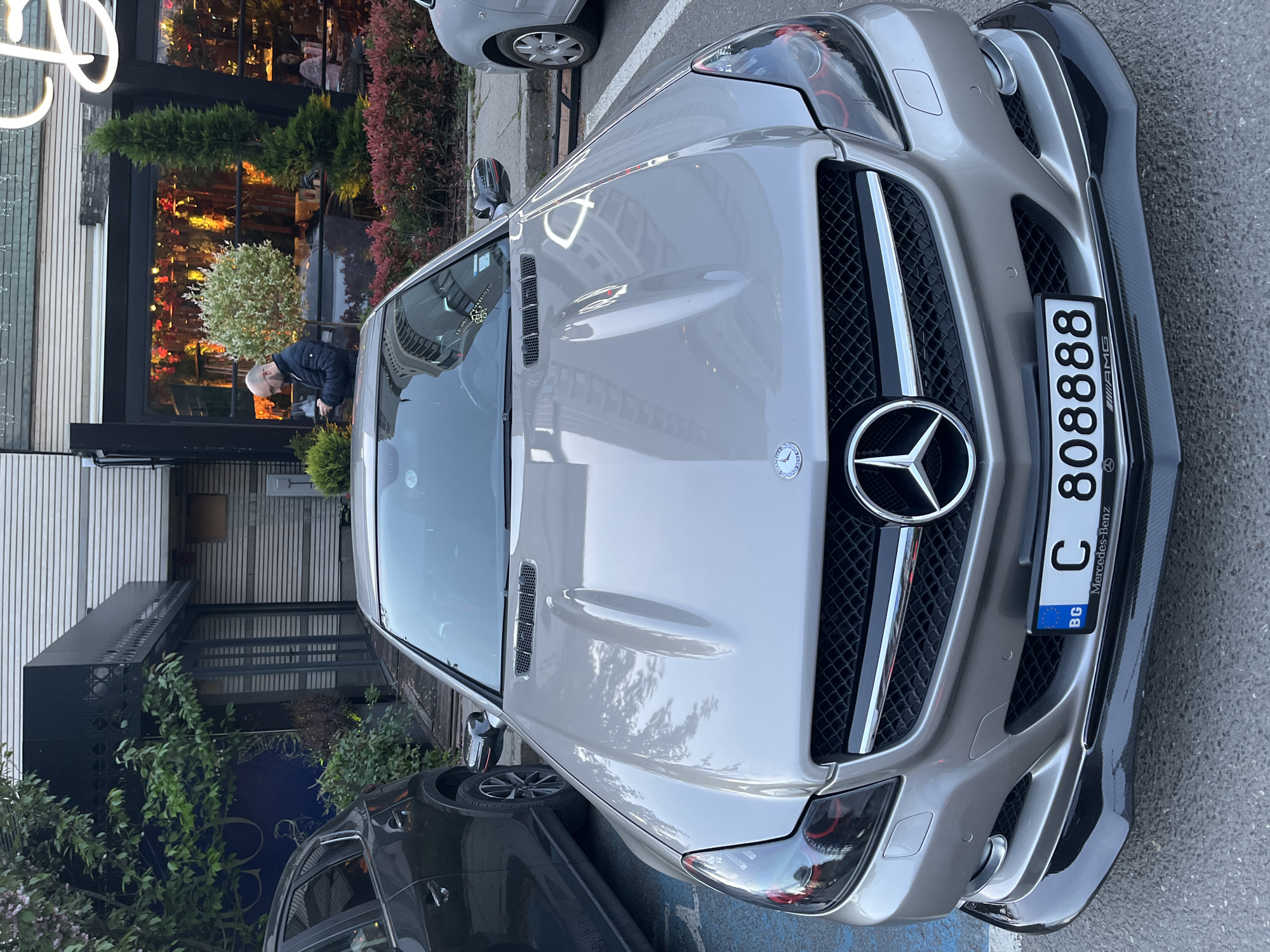 The image shows a silver Mercedes-Benz SL parked on a city street. The car has a Bulgarian license plate reading "C 808888." A person is bent over, possibly looking at the car. In the background is a building with plants and a storefront or restaurant. The image was most likely taken in Sofia, Bulgaria, based on the license plate.