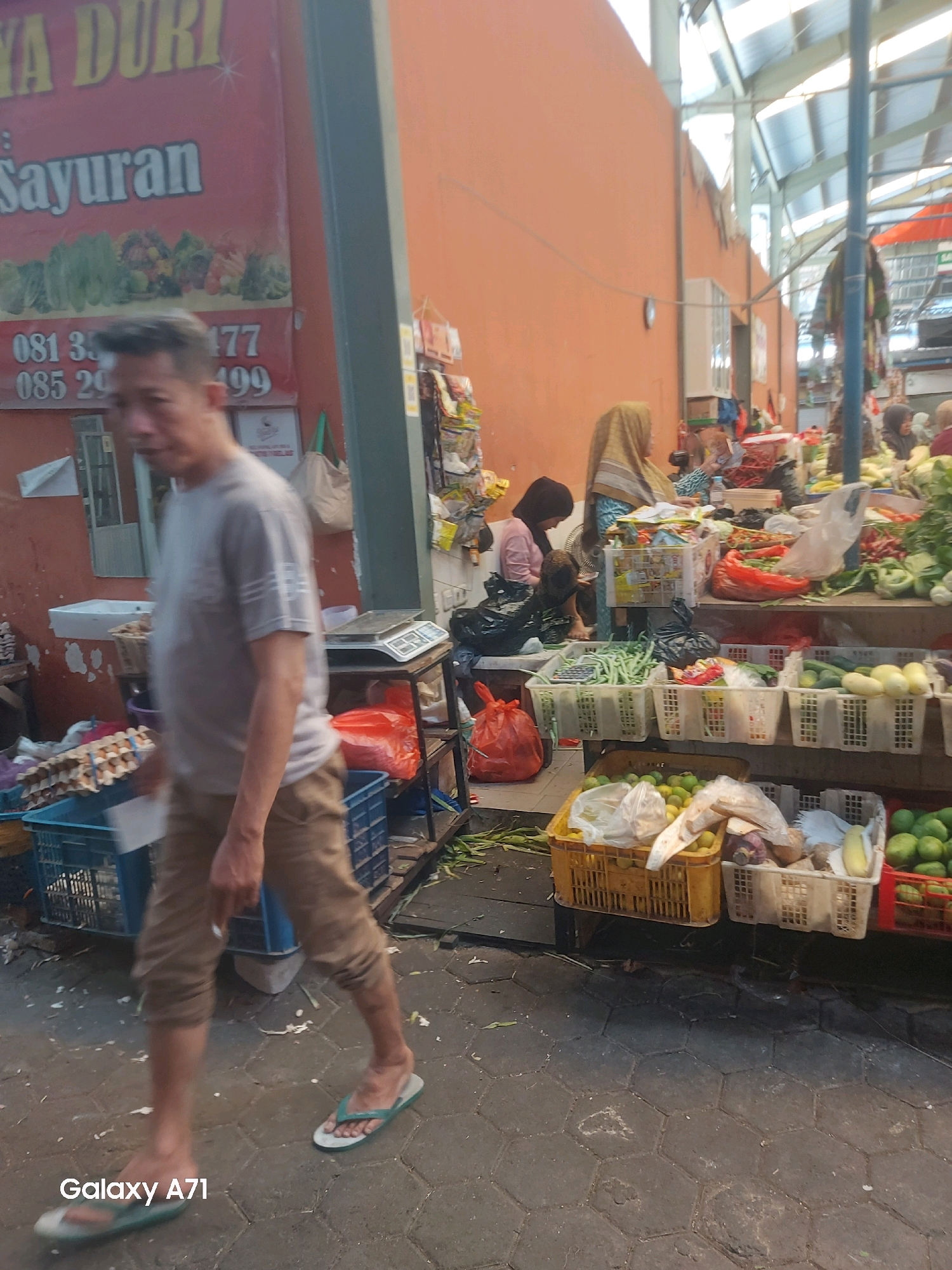 The image captures a bustling street market scene in Makassar, Indonesia. In the foreground, a man in a grey t-shirt and brown shorts walks on a paved walkway, wearing flip-flops. Behind him, a sign on an orange wall advertises "DURI SAYURAN" (Duri Vegetables) with contact numbers. To the right, several market stalls are visible, filled with a variety of fresh produce. Various baskets and crates overflow with green beans, cucumbers, squash, limes, and other fruits and vegetables. Shoppers and vendors, some wearing headscarves, are present among the stalls. The market appears to be covered by a translucent roof structure.
