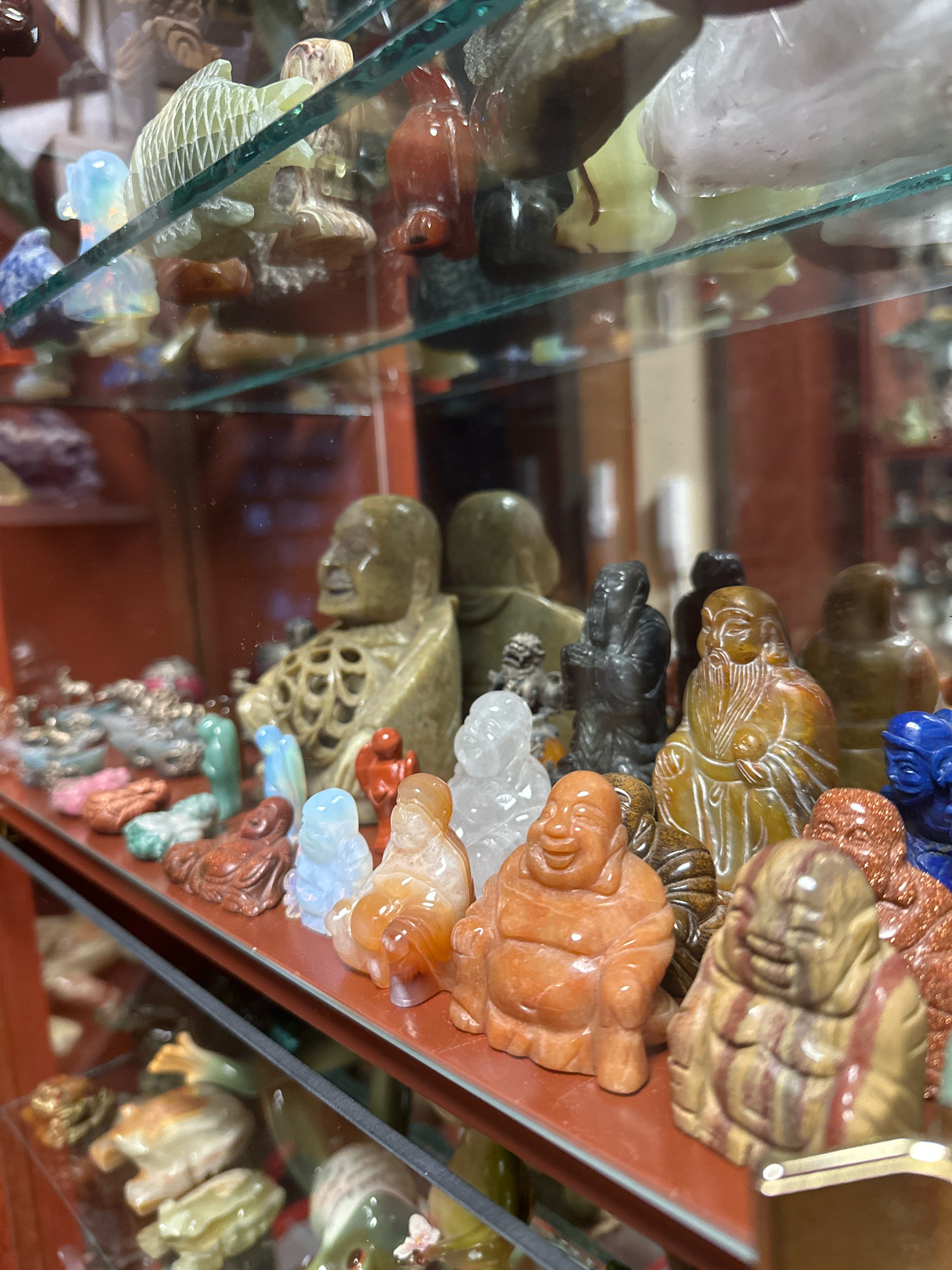 A close-up, angled shot captures a display of small, carved figurines made of various stones and minerals, arranged on glass shelves within a cabinet. The items appear to be of Asian origin, with many depicting Buddhist figures, animals, and mythical creatures. In the foreground, several plump, smiling figures resembling the Budai, or "Laughing Buddha," are prominently displayed. Some are crafted from translucent, orange-hued stone, while others are a mottled brown and red. Behind them, a row of darker, more intricately carved figures, including one with a long beard and another in a hooded robe, are visible. Further back, a larger, light-brown stone carving of a figure with a hollowed-out chest sits on the shelf. The background is blurred but suggests a retail or decorative setting, with hints of red and brown surfaces. The lighting is diffuse, highlighting the smooth textures and varied colors of the stone carvings. There is no indication of the city of Murmansk, Russia, within the visual information provided.