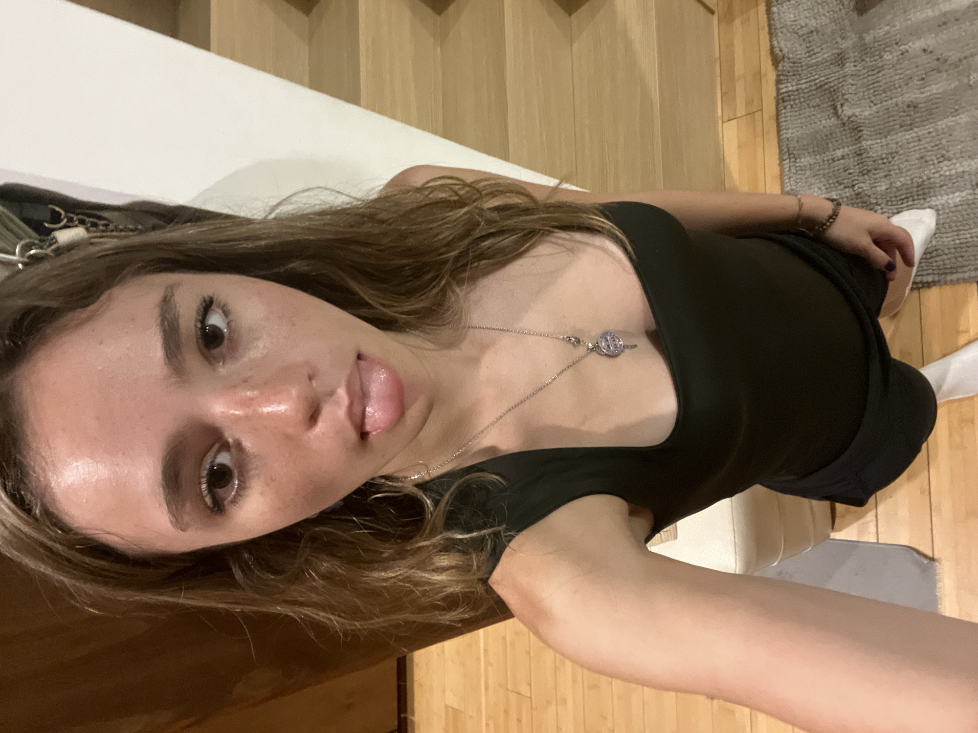 A young woman with light brown hair is positioned in the foreground, wearing a black top and a silver necklace. She has her tongue slightly extended. Her right arm extends toward the camera. She appears to be sitting on a white surface, possibly a bench. Behind her is a hardwood floor that transitions into a gray rug in the upper right corner. Above the floor are wooden stairs. The image appears to be taken indoors. The city of San Bernardino Tlaxcalancingo, Mexico is not visibly identifiable in the image.