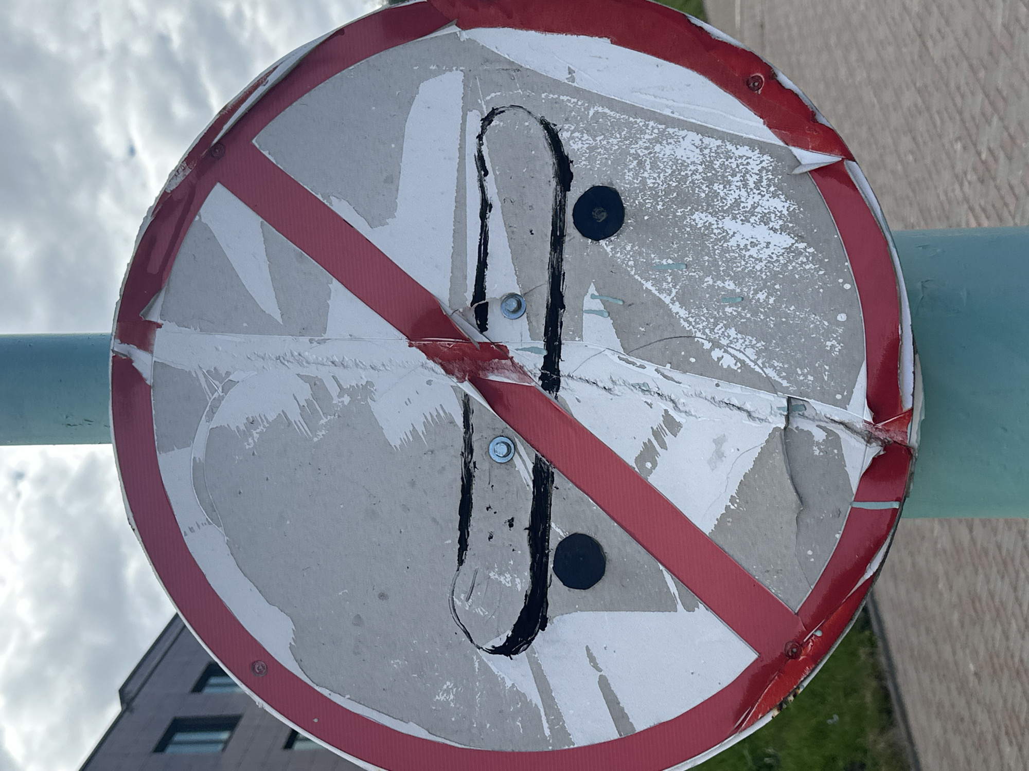 The image shows a close-up, slightly angled view of a damaged, circular traffic sign. The sign has a red border and a diagonal red stripe across it, indicating a prohibition. The white surface of the sign is heavily worn, with large sections of the reflective material peeling off, revealing a gray backing. Two black, roughly drawn vertical lines with curved bottoms are overlaid on the sign, resembling a stylized 'U' or a broken pair of parentheses. There are also two black circular stickers, one above and one below the drawn lines. The sign is mounted on a light blue metal pole or railing. In the background, a modern building with visible windows and a tiled facade is partially visible, along with a cloudy sky. The lighting suggests it is daytime. The condition of the sign implies it has been exposed to the elements and possibly vandalism.