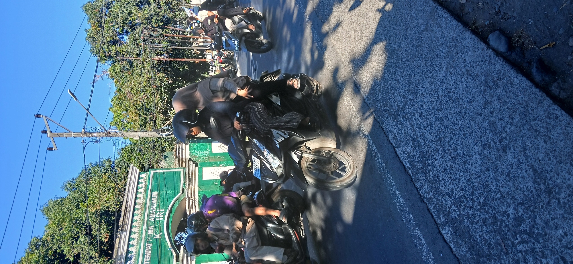 The image shows a street scene in Kediri, Indonesia, with several people riding motorcycles. The weather is sunny and clear, with a bright blue sky. In the background, there is a green building with text that appears to read "TEMPAT PEMAKAMAN UMUM KEDIRI". There are also trees and utility poles with power lines. The foreground shows a busy street with motorcycles in motion, casting shadows on the asphalt. One motorcycle in the foreground has two people riding on it, both wearing helmets. The person in the front is driving, and the person in the back is a passenger. Another motorcycle to the left has one person riding it, also wearing a helmet. Further down the street, other motorcycles and riders can be seen. The overall scene depicts a typical day on the road in an Indonesian city.