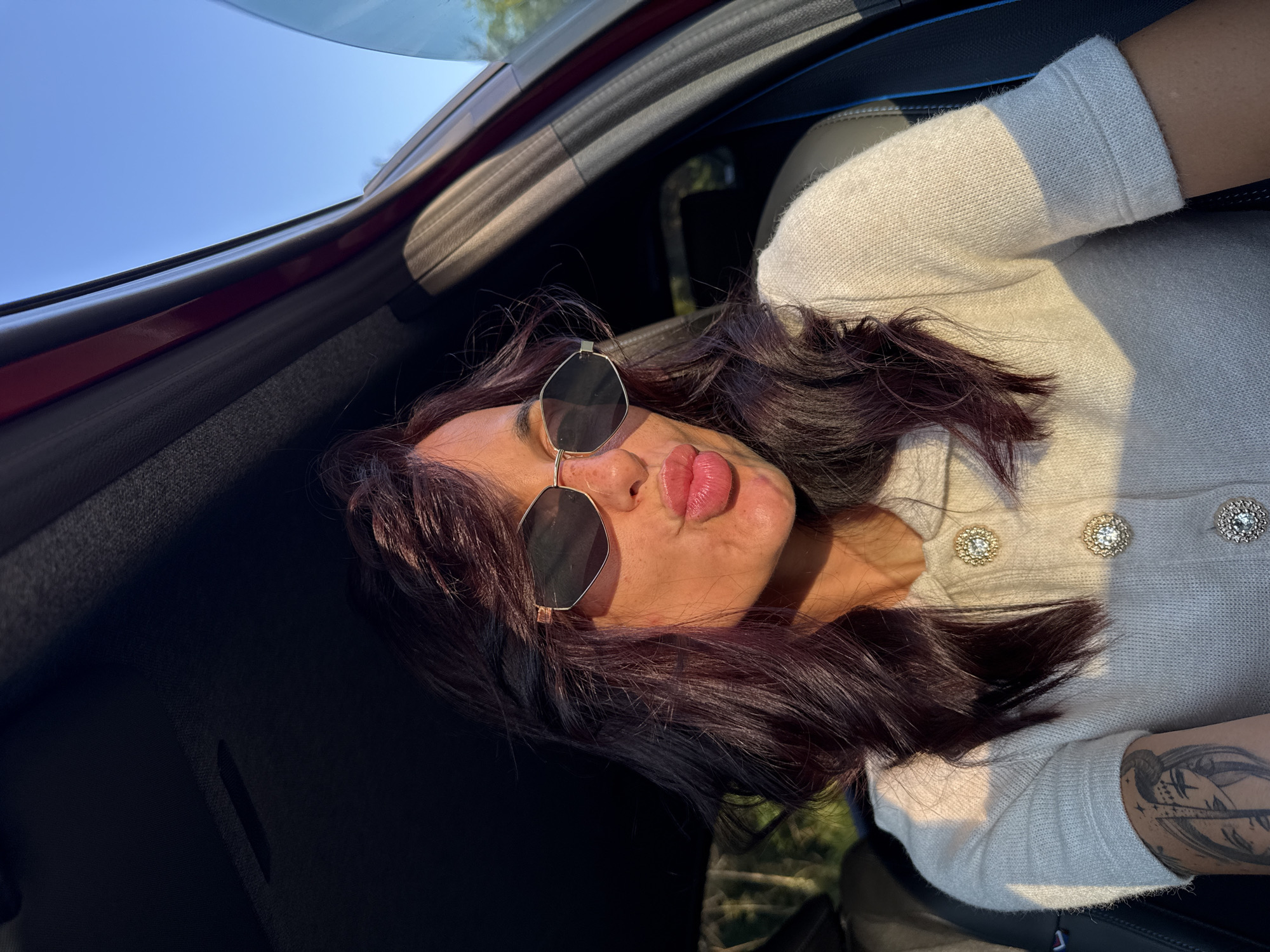 A woman with dark hair and sunglasses is sitting in the driver's seat of a car, leaning towards the camera with her lips puckered. The car's interior is visible, including the dashboard and a red trim on the door. The exterior shows a clear blue sky and greenery. The woman is wearing a light-colored sweater with decorative buttons. A tattoo is visible on her left arm. The scene appears to be illuminated by warm, late afternoon sunlight. The location is not specified within the image or the provided context.