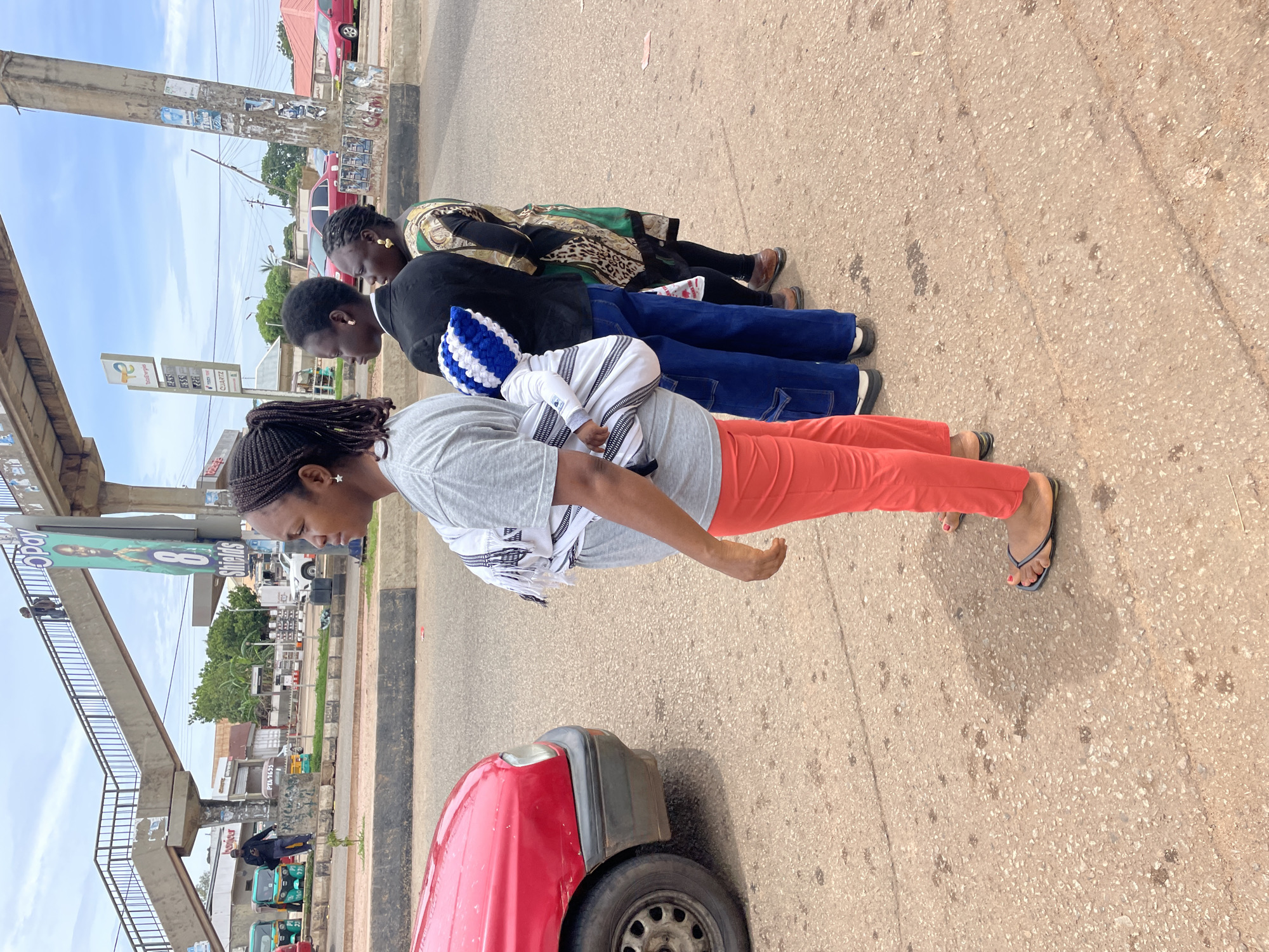 The image depicts a street scene in Gura Top, Nigeria. In the foreground, a woman wearing orange pants and a grey t-shirt is standing, with a baby wrapped in a white and black patterned cloth on her back. She is wearing flip-flops. To her right, a person in a black blazer and blue jeans stands, also with a baby wrapped similarly. Behind them, another woman is visible, wearing a patterned top and black pants.
In the background, a red car is parked to the left. Further back, a pedestrian bridge spans across the street, with people visible on it. A sign with a man's face and the word "Shields" is posted on a pole supporting the bridge. Other signs, including a "Total Energies" sign with fuel prices, and various buildings and trees are visible along the roadside. The ground is paved with asphalt, showing signs of wear and tear. The sky is cloudy.