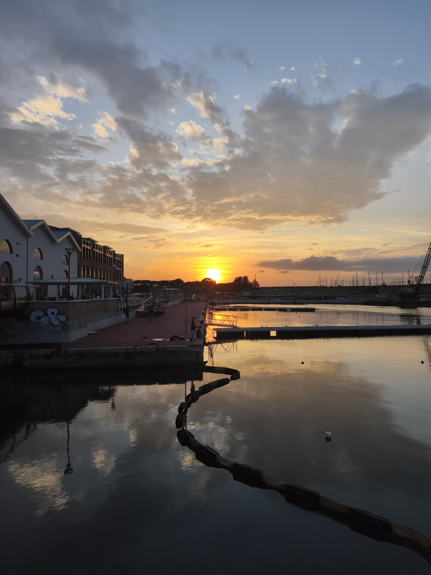 This image depicts a sunset over a harbor in Livorno, Italy. The sun is low on the horizon, casting a warm orange and yellow glow across the sky and reflecting on the calm water. In the foreground, the water is dark, with reflections of the sky and clouds visible. A dark, segmented boom floats on the water's surface.
Along the left side of the image, there is a row of modern buildings with white facades and dark roofs. These buildings appear to be shops or restaurants, with windows and covered outdoor seating areas. There are a few people visible on the paved walkway in front of these buildings. Graffiti is spray-painted on a wall in the lower left section of the image.
Further into the harbor, there are docks and numerous sailboats moored. A large crane is visible on the right side of the harbor, suggesting ongoing construction or maintenance. The water's surface in the midground and background is smoother, with clearer reflections of the sky, boats, and structures.