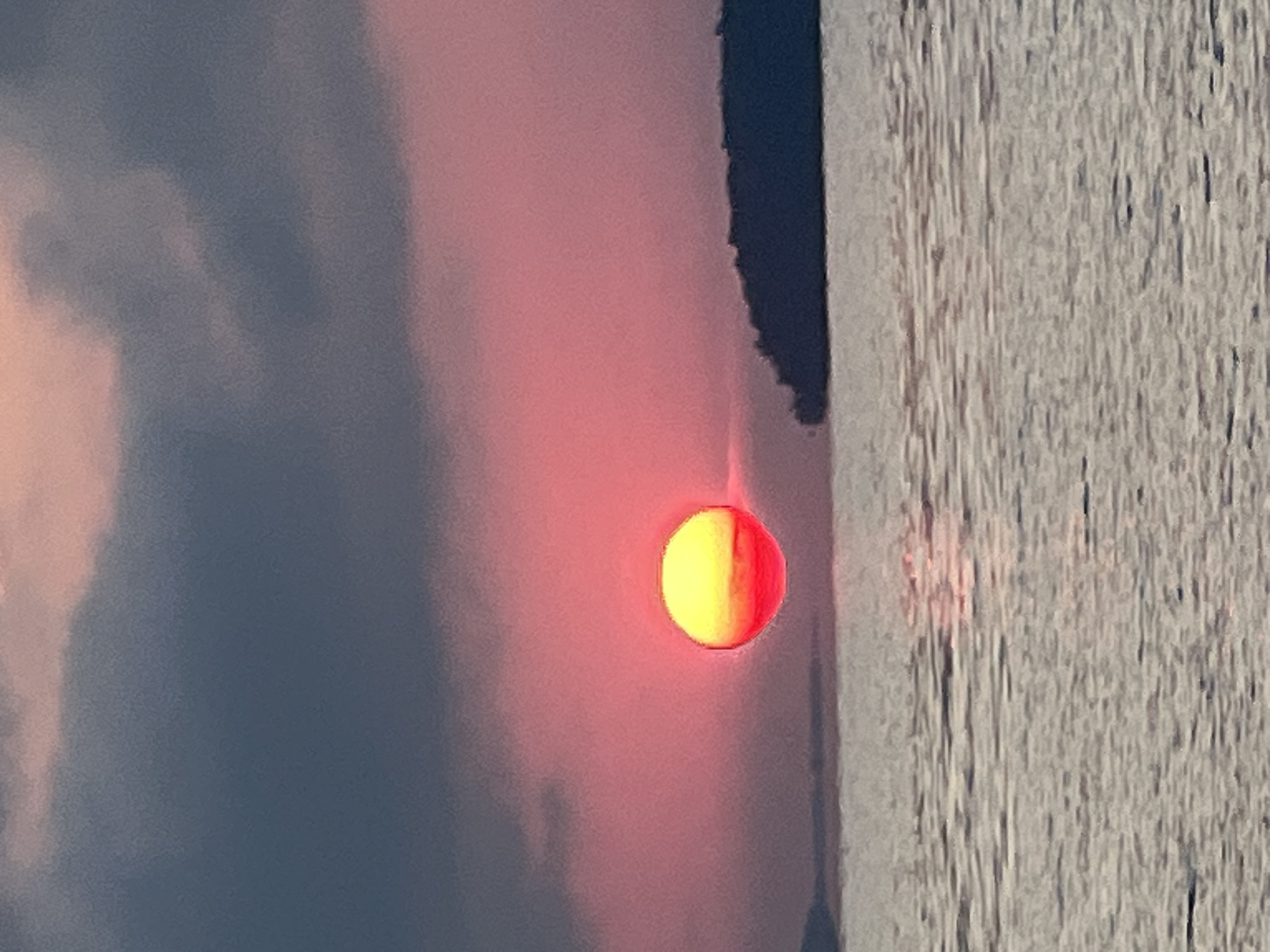 A low-angle, medium shot captures a sunset over a body of water. The sun, a bright orb of yellow and red, is partially obscured by dark, hazy clouds that cover the upper portion of the frame. The sky transitions from a deep blue-gray at the top to a vibrant pink and orange closer to the horizon. In the mid-ground, a silhouetted island with a jagged coastline juts out from the right side of the frame. The foreground is dominated by the rippling surface of the water, reflecting the colors of the sunset. The scene appears to be set in Novopichugovo, Russia, possibly on the shore of Lake Baikal. No people are visible in the image.