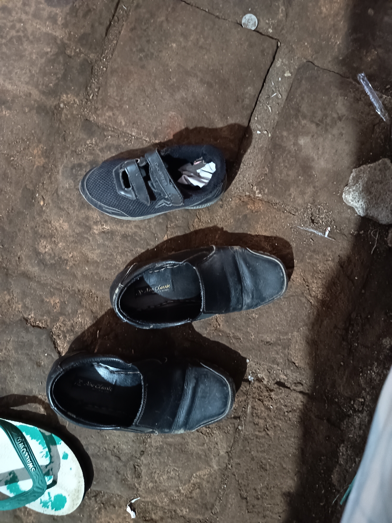 The image shows several footwear items on a dirty, cracked stone or concrete surface, possibly a pathway or ground. From top to bottom, there is a black sneaker with velcro straps, a black leather dress shoe, another black leather dress shoe, and a flip-flop with a white sole and green and white patterned straps. The overall scene suggests an outdoor or neglected environment, possibly in a less developed area. The lighting appears to be artificial and low, casting shadows. There are also small pieces of debris scattered on the ground, including what might be a coin and a piece of plastic. No people are directly visible in the image, but the footwear implies their recent presence or absence. The location is stated to be Krampilan, Indonesia, which provides a geographical context for the scene.