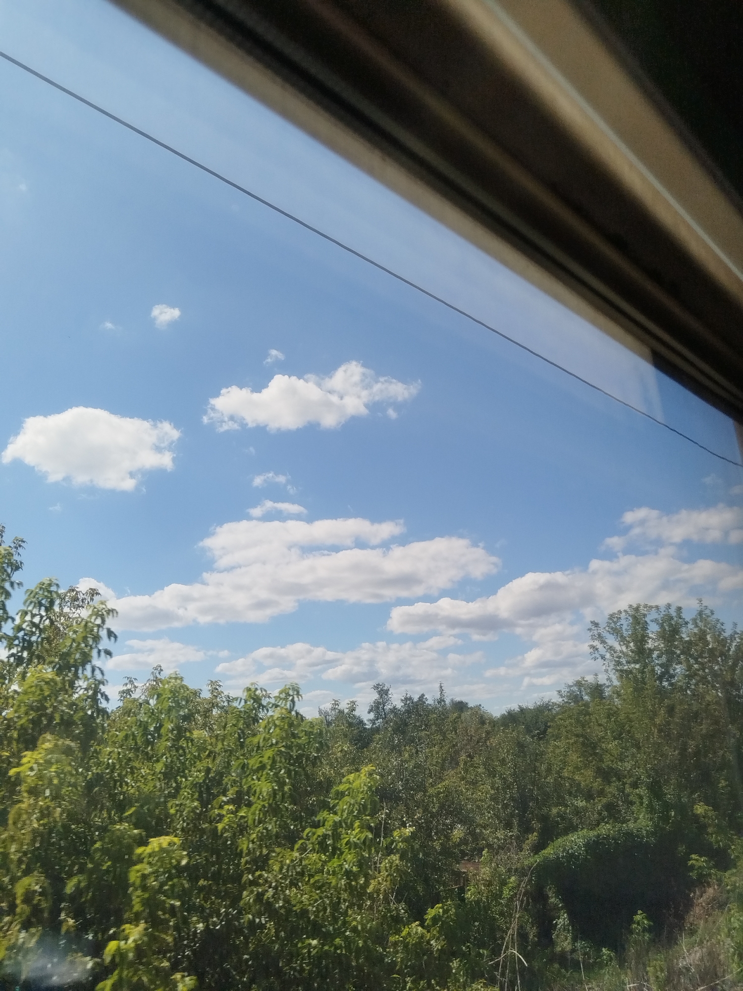 The image depicts a view from a window overlooking a landscape of green trees and a blue sky with white clouds. The top of the frame is partially obstructed by a dark, textured frame, possibly the interior of a vehicle. A thin, dark line runs diagonally across the sky. The scene appears to be a natural outdoor environment, potentially rural or suburban, with no visible buildings or people. The light suggests it is daytime. The location is not specified within the image itself, and therefore cannot be determined as Yumatovo, Russia.